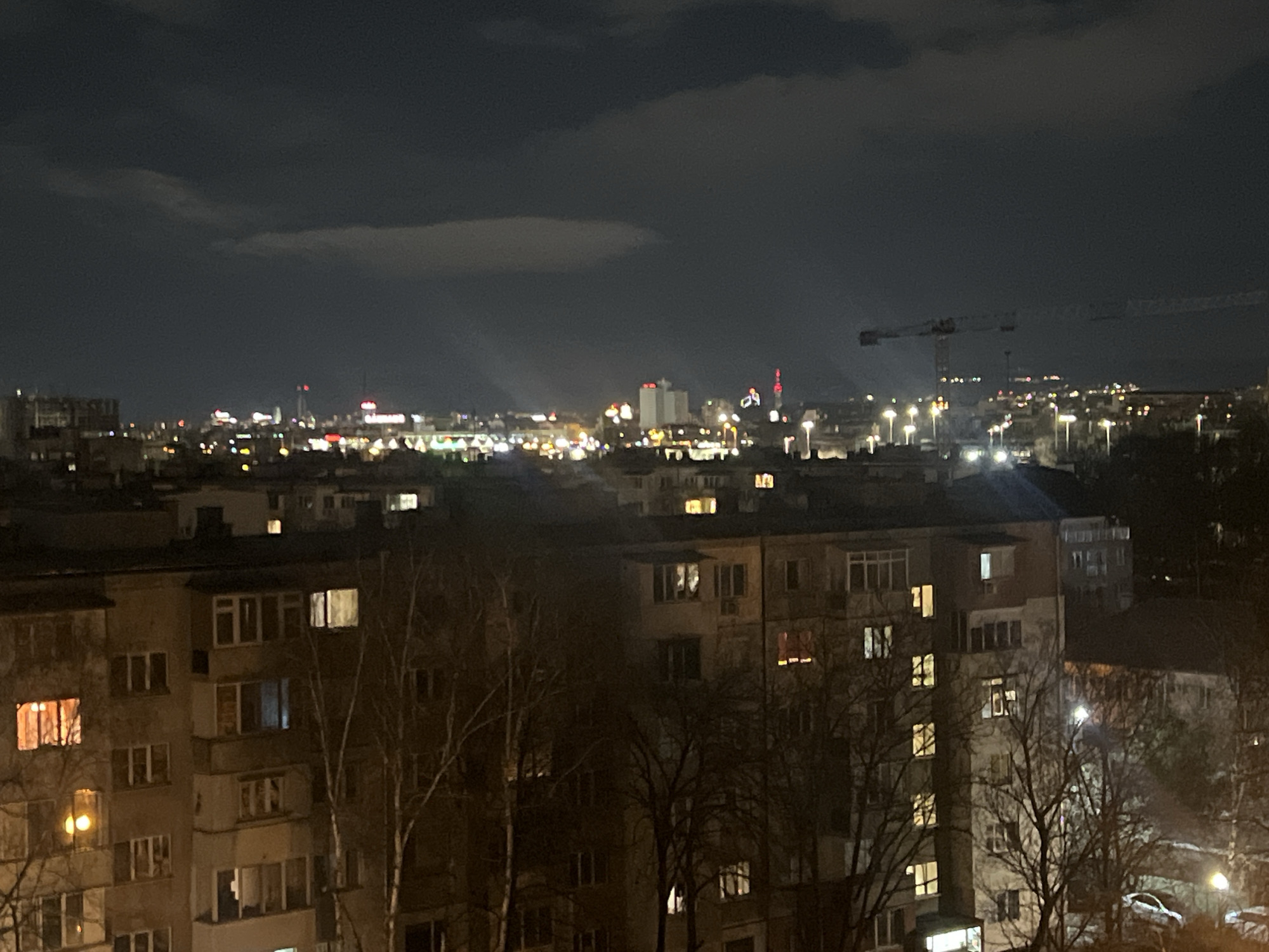 The image shows a nighttime cityscape in Sofia, Bulgaria. In the foreground, are residential buildings with illuminated windows, partially obscured by leafless trees. In the background, the city skyline is visible, with various buildings and structures illuminated. A crane is prominent in the distance. The sky is dark and cloudy, with lighter patches suggesting the presence of city lights.