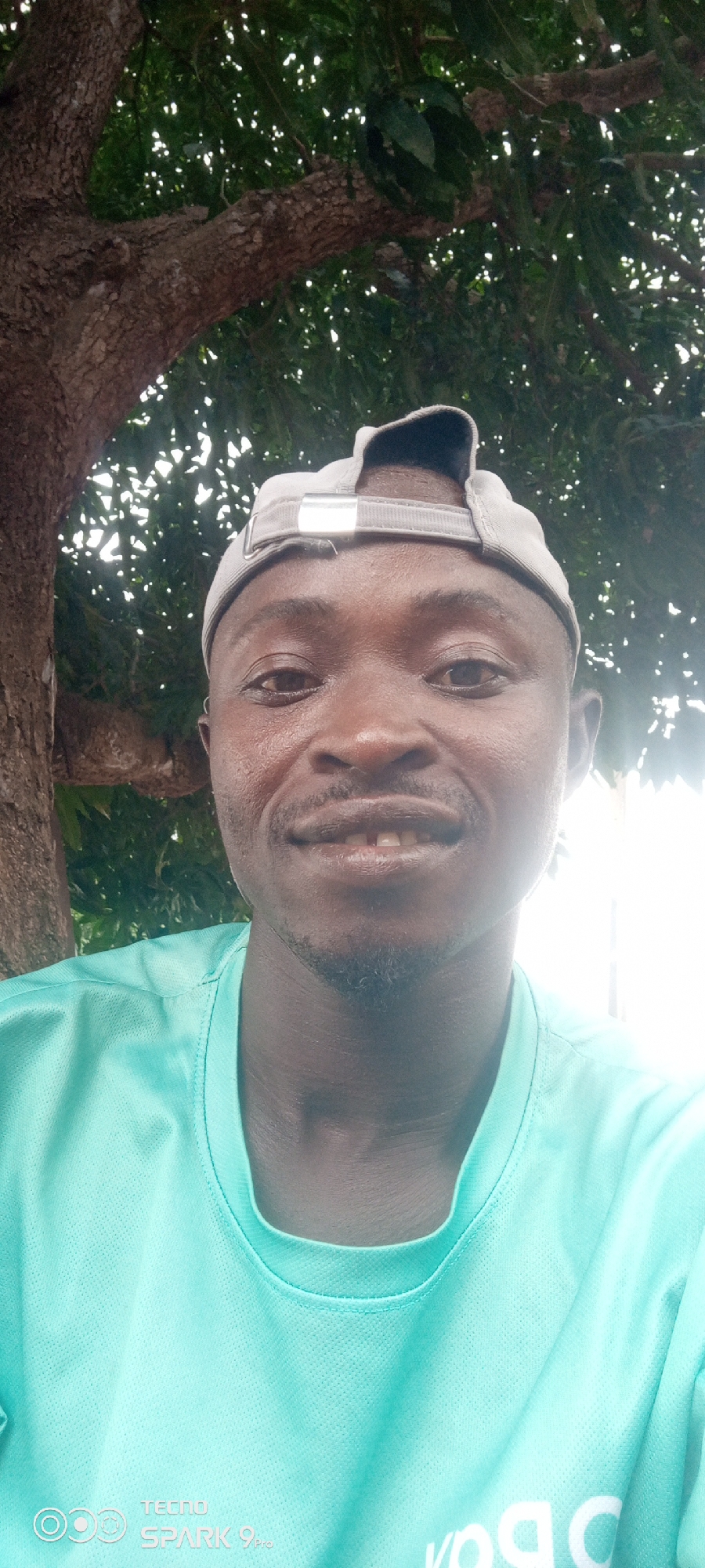 A selfie shows a Black man wearing a light gray baseball cap and a teal t-shirt, smiling at the camera. He has a light beard and a gap between his front teeth. The man is outdoors, with a large tree and lush green foliage in the background. The lighting suggests it is daytime, with sunlight filtering through the leaves. The bottom of the image shows a watermark with "TECNO SPARK 9 Pro" branding. The location is identified as Kurmin Dangana, Nigeria.