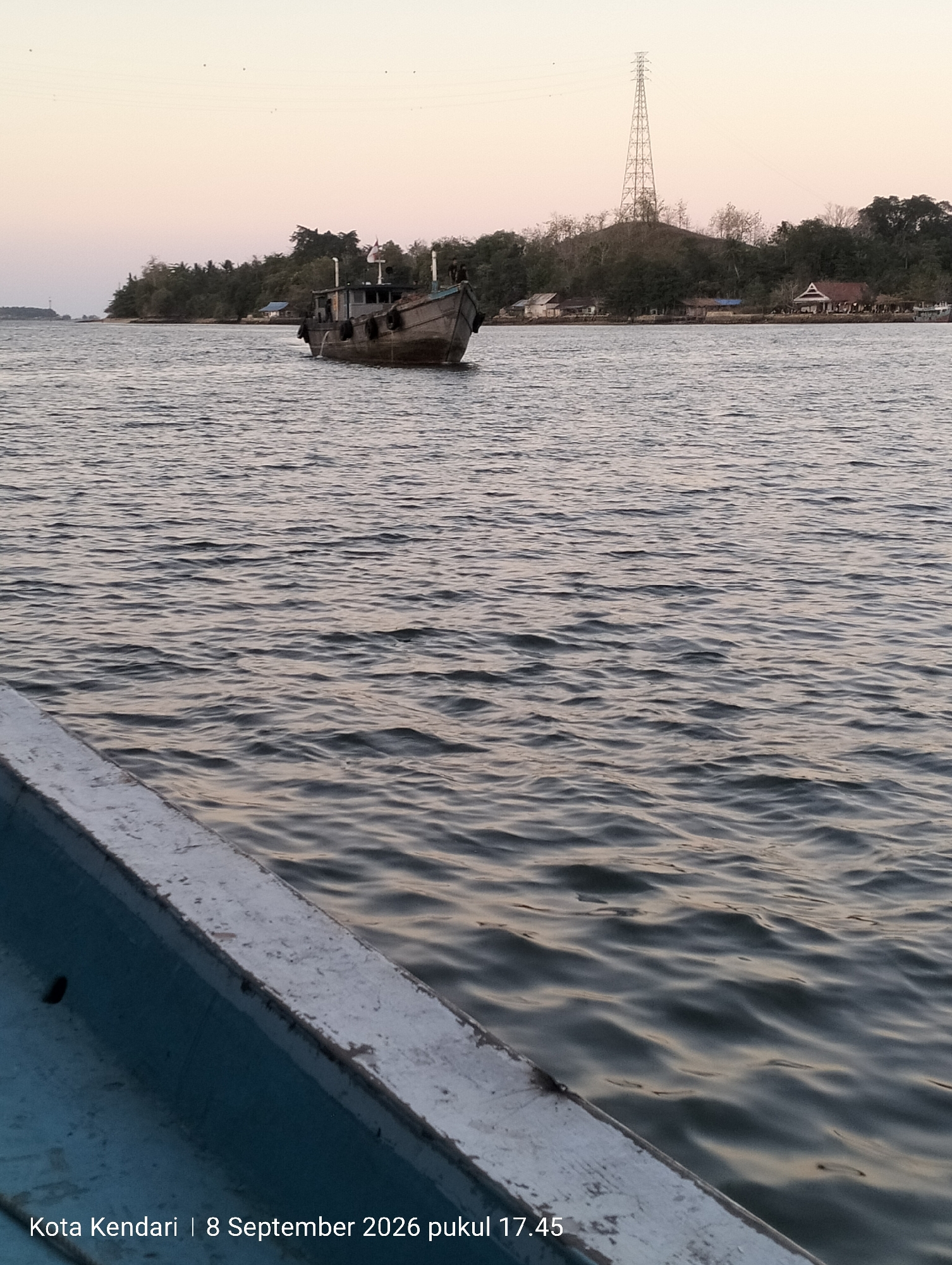 The image shows a view from a boat on the water, looking towards a coastline in Kendari, Indonesia. In the mid-ground, a single, weathered barge with visible ropes and mooring lines is positioned on the water, appearing stationary. The barge is dark in color and shows signs of wear. Behind the barge, the coastline is characterized by a dense line of trees and vegetation. Scattered among the trees are several buildings, including houses with traditional pitched roofs and what appears to be a residential area. Prominently featured on a hill behind the settlement is a tall, lattice-style transmission tower. The sky above is a soft gradient of pale pink and orange, suggesting either dawn or dusk. The water in the foreground exhibits small ripples and reflects the muted colors of the sky. The lower left corner of the image displays a portion of the boat from which the photograph is taken, showing its blue interior and a weathered white gunwale. Text overlaid at the bottom of the image reads "Kota Kendari | 8 September 2026 pukul 17.45", indicating the location and timestamp.