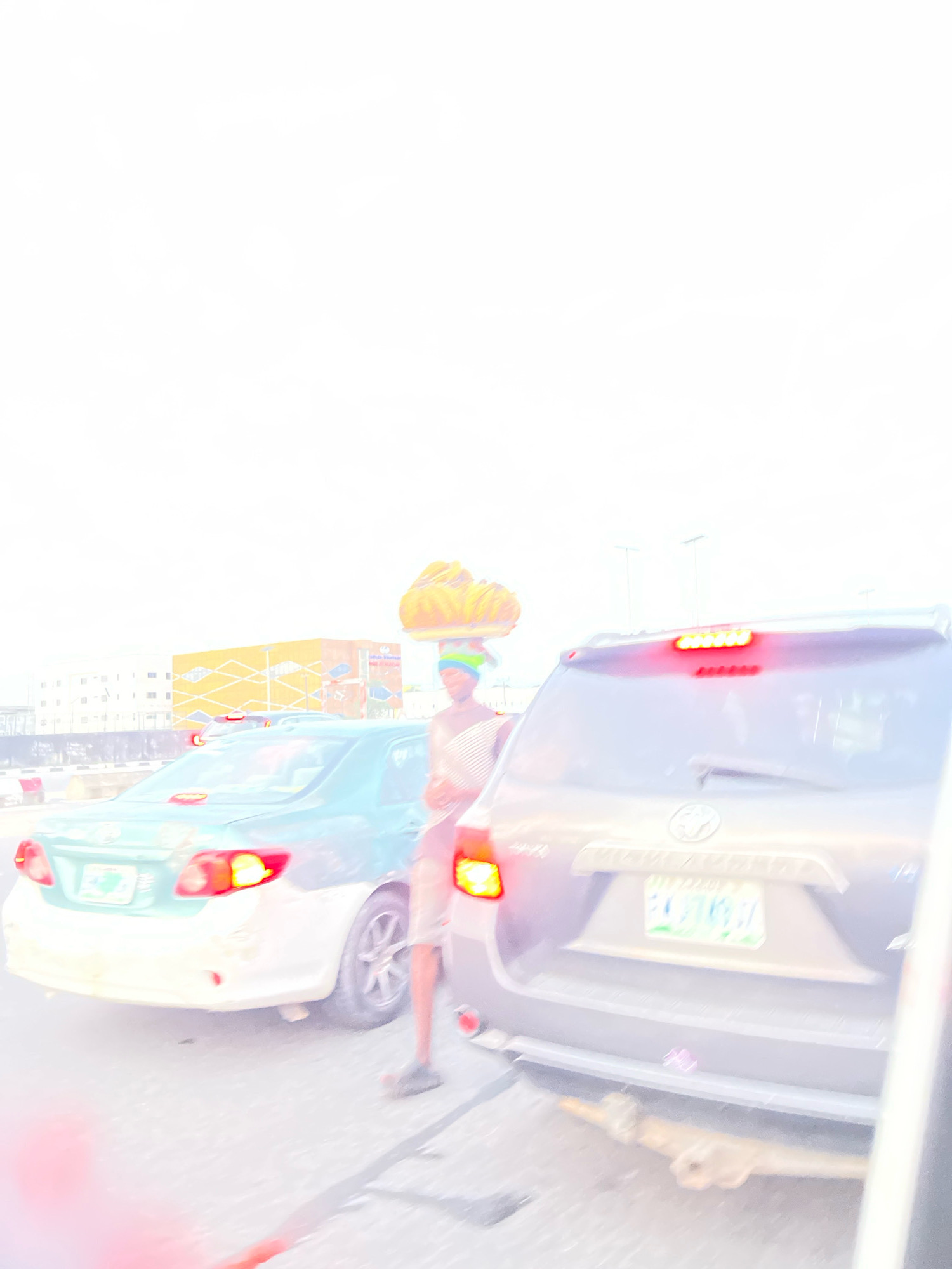 The image depicts a street scene in Lekki Peninsula II, Nigeria, characterized by a traffic jam. A person is standing between two cars, balancing a basket of yellow fruits, likely bananas, on their head. The person is wearing a blue and green patterned headwrap and a light-colored garment. To the left, a light blue car with a white lower body is visible, its brake lights illuminated. To the right, the rear of a darker colored SUV is shown, also with its brake lights lit. The SUV has a license plate. In the background, a building with a yellow and orange facade is present, along with other structures and utility poles. The sky is bright and overcast. The overall impression is one of a typical urban traffic situation.