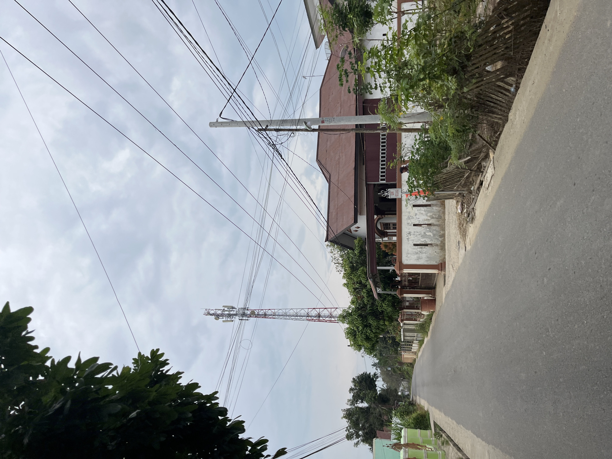 A photograph taken from the perspective of standing on an asphalt road in the city of Tanahterban, Indonesia, shows a street lined with houses and a telecommunications tower in the distance. The sky is overcast with a soft gray light. On the left side of the road, a green gate and part of a house are visible, with lush green foliage dominating the foreground. The right side of the road features a white house with a reddish-brown tiled roof, a white fence, and more vegetation. Several power lines crisscross the sky, supported by a utility pole in the mid-ground. The telecommunications tower, a prominent structure, stands tall in the background, with its red and white frame and antennas clearly discernible. A small section of a bamboo fence runs along the side of the road, overgrown with green plants. A person is partially visible in the background, near the white house.