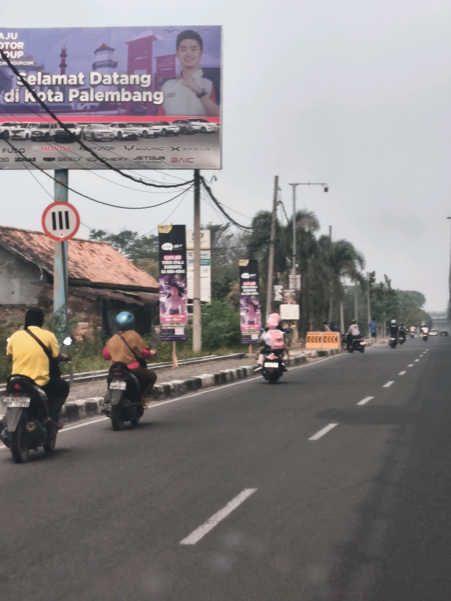 The image depicts a street scene in Palembang, Indonesia, featuring a billboard advertising "Selamat Datang di Kota Palembang" (Welcome to Palembang City) with images of cars and a smiling man. Several motorbikes are present on the road, some with riders. One motorbike in the foreground is ridden by a person wearing a yellow shirt. To the right, another motorbike carries a rider in an orange jacket and a blue helmet. Further down the road, more motorbikes are visible, with some individuals standing or walking. The environment includes a road with lane markings, sidewalks, trees, and buildings. A traffic sign with the number "III" inside a circle is also visible. The sky is overcast.