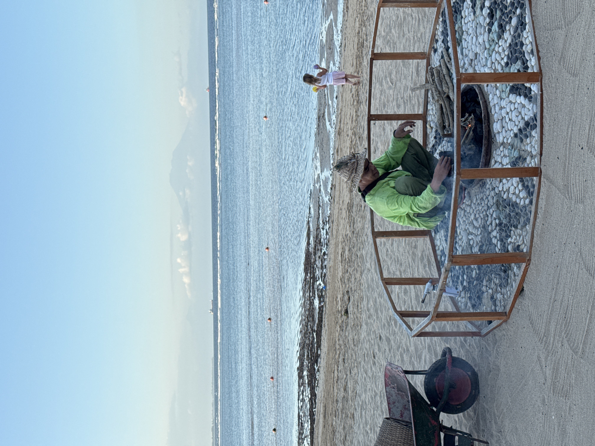 A man in a straw hat and a bright green long-sleeved shirt crouches in a hexagonal fire pit on a sandy beach. The fire pit is bordered by wooden posts and filled with a mosaic of light and dark grey stones. The man is tending to a small fire in the center of the pit, surrounded by a pile of logs. To the left of the fire pit, a red and green wheelbarrow is parked on the sand, partially filled with a woven basket. In the background, the blue ocean stretches to the horizon, with white waves breaking on the shore. A young girl in a white tank top and pink shorts stands in the shallow water, holding yellow and purple toys. The sky is a clear, pale blue with a faint outline of a mountain range in the distance. This scene is located on the beach in Kuta Selatan, Bali, Indonesia.
