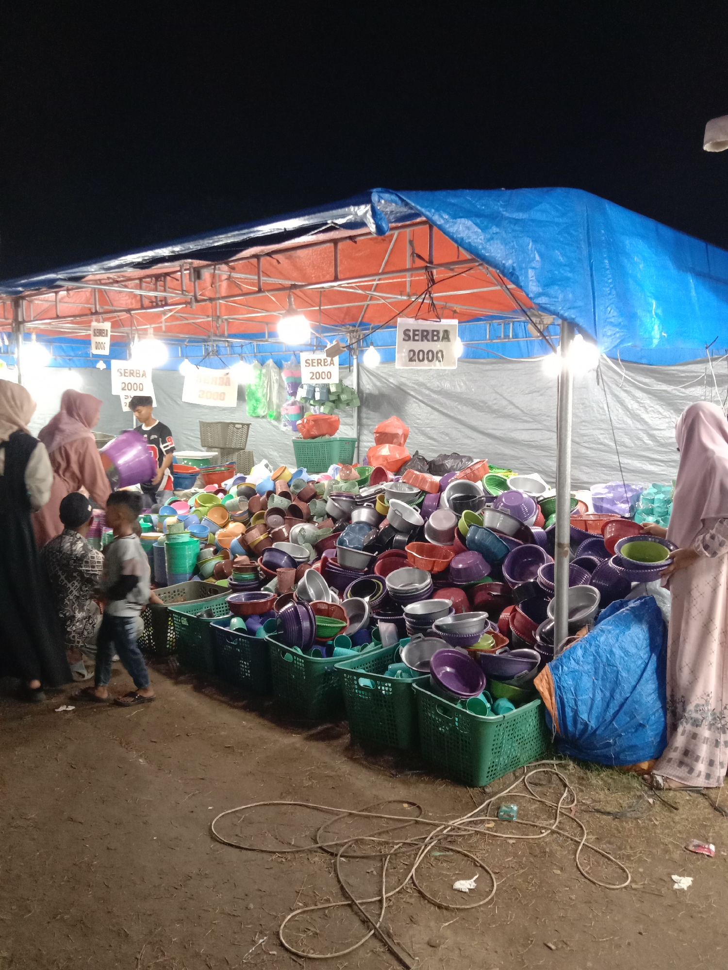 The image depicts a vendor stall at night, likely located in Geulumpangsulu Timu, Indonesia. The stall is covered by a blue and orange tarp, with signs that read "SERBA 2000". The stall contains a large collection of plastic bowls and containers, many of which are in green plastic crates. Several people are browsing the items. Two women wearing hijabs are visible on either side of the frame, inspecting the merchandise. A boy in a grey t-shirt and jeans is walking past the stall. A boy in a t-shirt is holding a purple bowl. Electrical cords are on the ground. The ground is dirt.