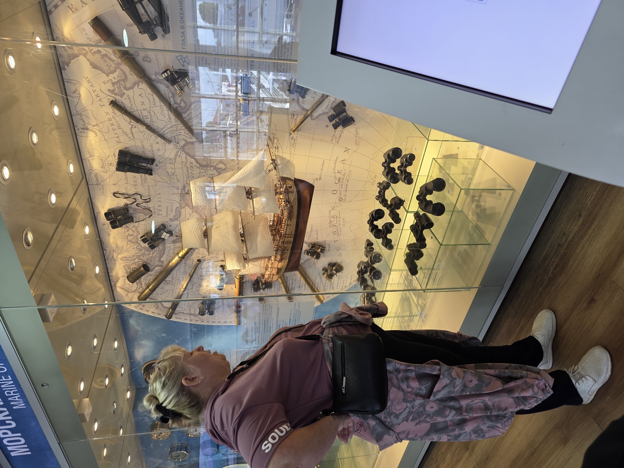 A woman stands in front of a museum display case in Kaliningrad, Russia. The display case contains a model of a sailing ship, antique navigational instruments like telescopes and binoculars, and maps. The woman, wearing a pink hoodie and black pants, is looking at the display. The museum exhibit appears to be related to maritime history.