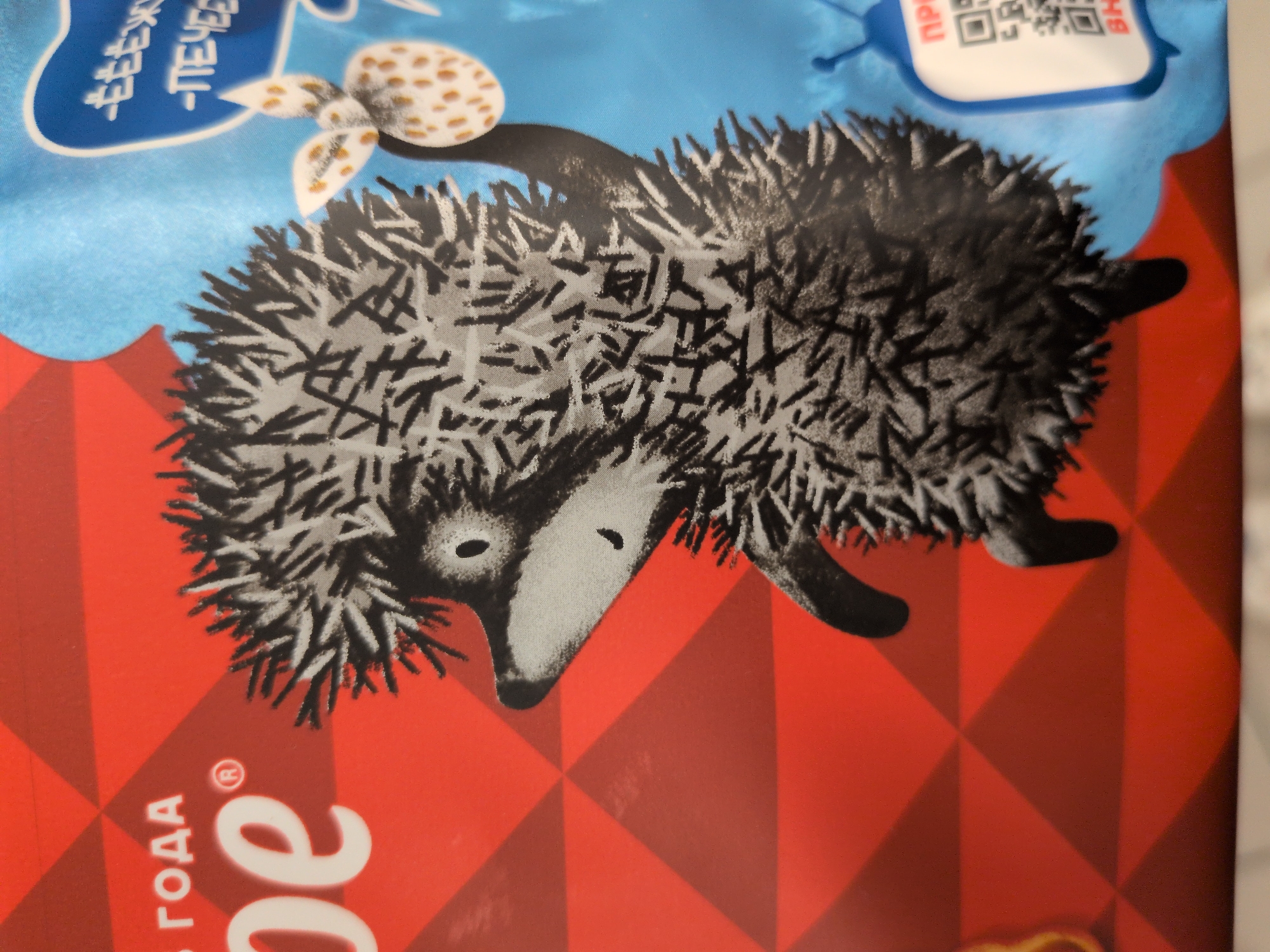 The image displays a vibrant product package, likely for cookies or biscuits, featuring an illustrated hedgehog. The hedgehog, depicted with black and grey quill-like spikes, a black snout, and black eyes, appears to be walking on a red checkered surface towards the left. Above the hedgehog, the background is a bright light blue, resembling a sky. Also in the blue area is a whimsical white, spotted, mushroom-like object with a small tail, possibly representing a berry or a specific type of cookie.
Notable details include the contrasting colors of the package: a light blue top half and a bold red checkered pattern on the bottom half. The illustration style is artistic and playful.
Visible text on the package, written in Russian, provides clues about the product. In the top left, white text on a blue background reads "ЁЁЖИК" (Yozhik - Hedgehog) and "-ПЕЧЕНЬЕ" (Pechenye - Cookies/Biscuits), indicating "Hedgehog Cookies." In the top right, a white speech bubble with a blue outline contains a black QR code and partially visible text "ПР" and "ВЫ", possibly related to a prize or contest ("ПРИЗ" - Prize, "ВЫИГРАЙ" - Win). In the bottom left, white text against the red background includes "ГОДА" (Goda - Year) and a large letter "е" with a registered trademark symbol (®), suggesting part of a brand name. The overall impression is a cheerful design for a children's snack.