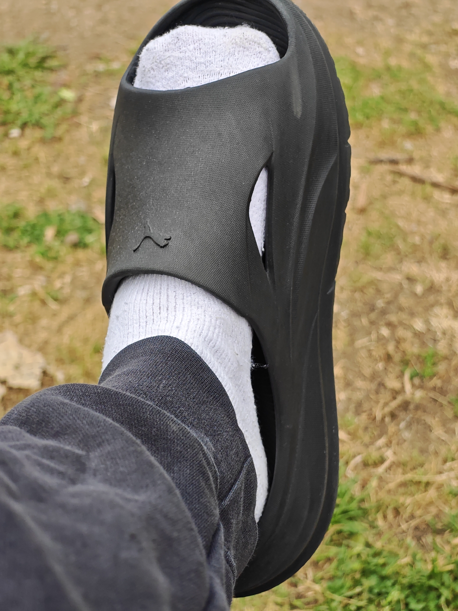 A close-up, outdoor, low-angle shot shows a person's left foot wearing a white sock and a black slide sandal. The sandal has a molded footbed and a wide strap with a cutout pattern. A small, dark logo, resembling a leaping feline, is visible on the sandal's strap. The foot is covered by a dark grey sweatpant. The background consists of dry grass and some green patches, suggesting an unpaved area. The lighting is natural and appears to be daylight. No specific location within Sofia, Bulgaria, can be identified from the visual information provided.