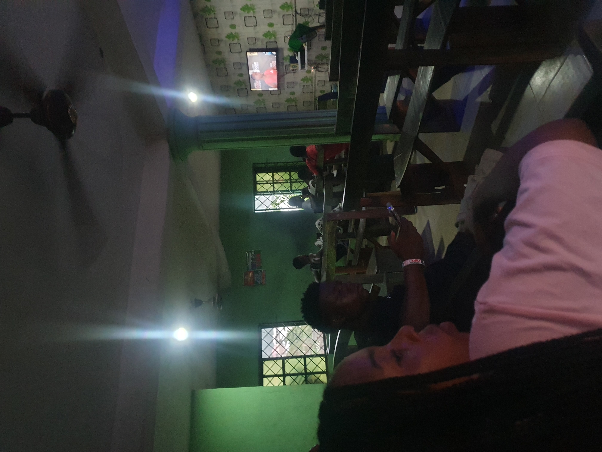 This image, taken in Lokoja, Nigeria, depicts an indoor gathering space, likely a casual restaurant or lounge, during the evening or night. The room features vibrant green walls and a ceiling adorned with patterned wallpaper containing light-colored squares.
In the foreground, a person with braided hair and a light pink top is visible, looking towards the left side of the frame. Their arm is also visible, holding a small, reflective object. In the midground, a man with short dark hair and a black shirt sits at a wooden table, gazing towards the right. He wears a wristband on his left wrist. Other indistinct figures are scattered throughout the space, occupying various wooden tables and chairs, suggesting a social environment.
The scene is dimly lit overall, relying on artificial light sources which create bright spots and lens flares, indicating the time of day. Two ceiling fans are visible, one prominently in the foreground and another partially cropped. Two windows with metal grilles are seen on the green wall, but the darkness outside confirms it is after sunset.
Mounted on the patterned ceiling wall, a flat-screen television displays what appears to be a sports match, possibly boxing or wrestling, featuring two figures. The content on the screen is a focal point for some in the room. A poster or sign is affixed to the green wall near the window, but its text is illegible. The atmosphere appears relaxed, with people socializing and engaging with the television content.