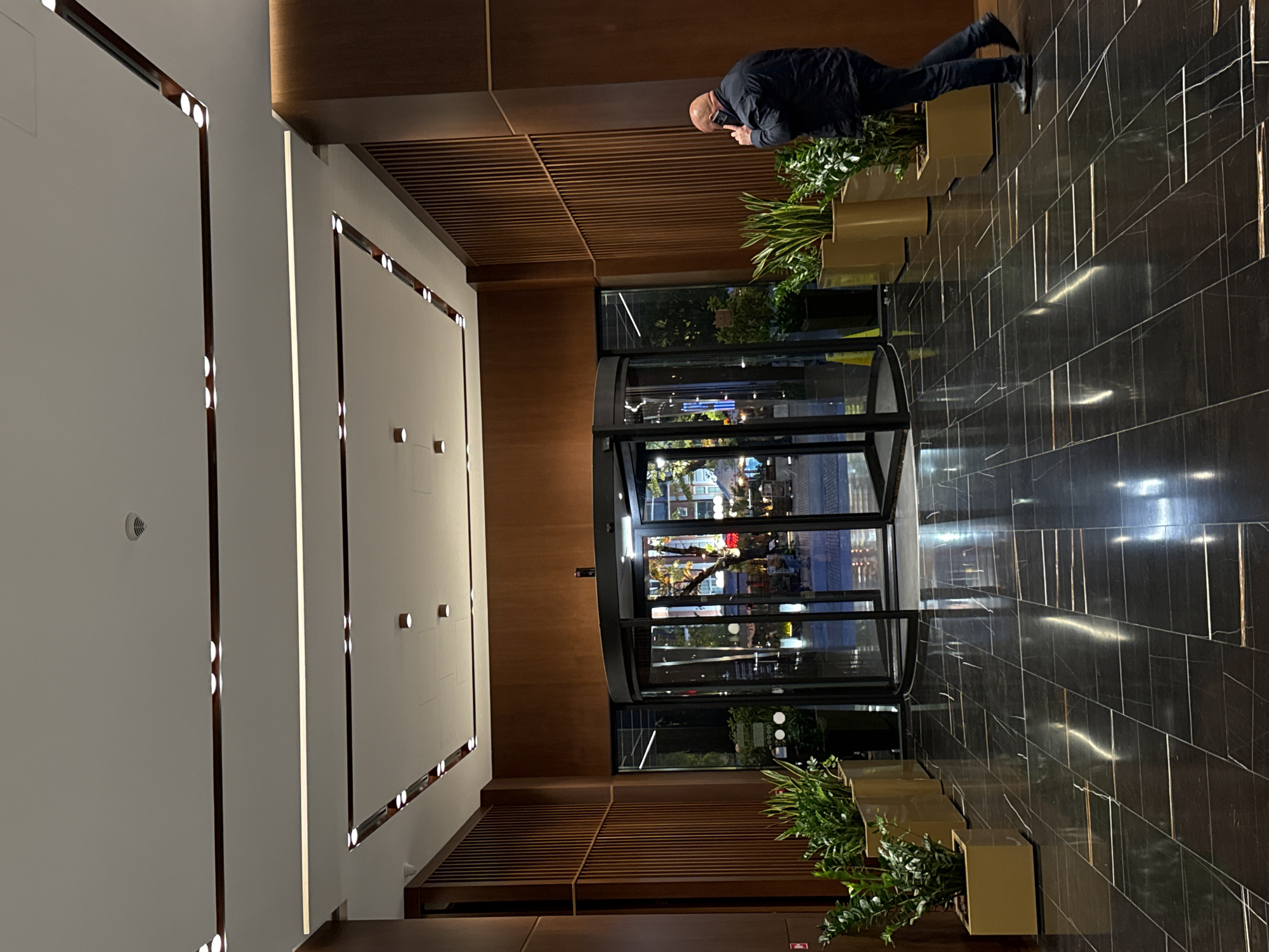 A photo taken in Varna, Bulgaria shows the entrance to a building, likely a hotel or business, with a revolving door. A man stands nearby, looking at his phone. Planters with green foliage flank the doorway. The interior of the building appears brightly lit. The floor is dark marble with light veining, and the walls are a combination of light plaster and dark wood.