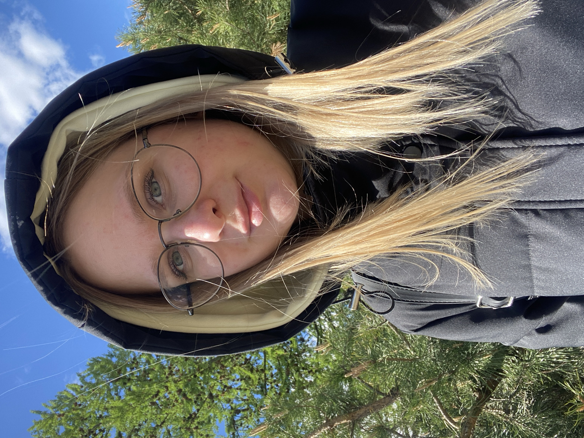 A young, light-skinned person with blonde hair and round eyeglasses is centered in the frame. The person is wearing a black hooded jacket with a light-colored lining and is looking towards the camera. The background consists of green pine tree branches and patches of blue sky with light clouds. The location cannot be determined to be Izhevsk, Russia based on the visual information provided.