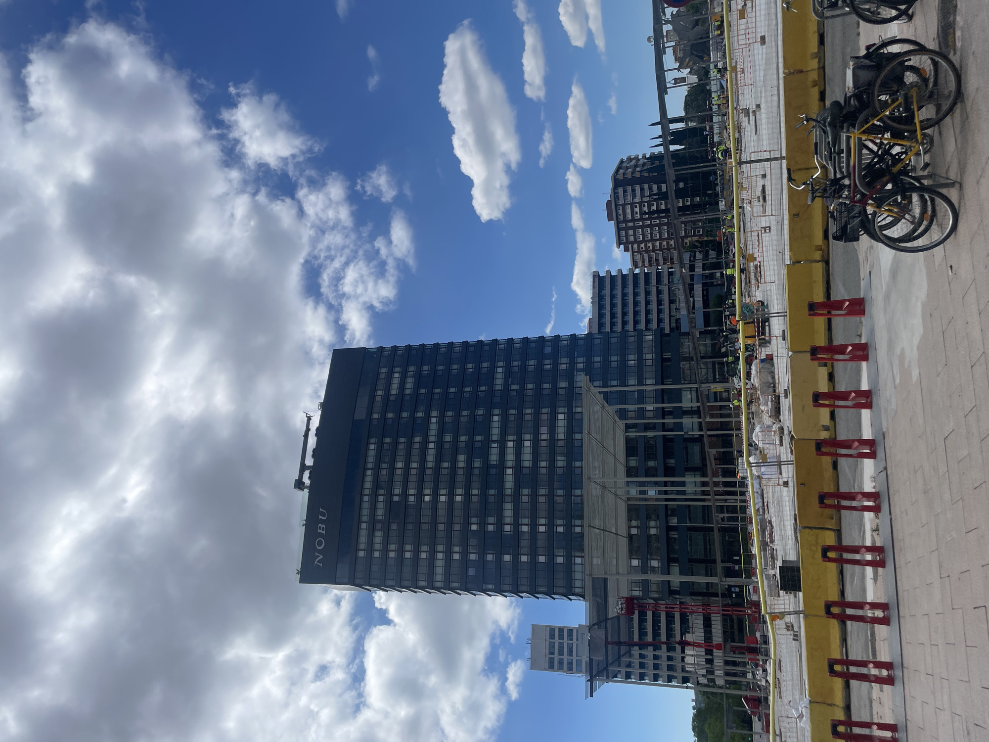 The image depicts a view of the Nobu Hotel in Barcelona, Spain. The building is a high-rise structure with a dark facade and numerous windows. The name "NOBU" is visible on the side of the building. The sky is blue with scattered white clouds. In the foreground, there's a construction site barrier. Bicycles are parked alongside the construction barrier. Some workers are present at the location. The surrounding area includes other high-rise buildings, indicating an urban environment. The photograph is taken at a low angle, emphasizing the height of the building.