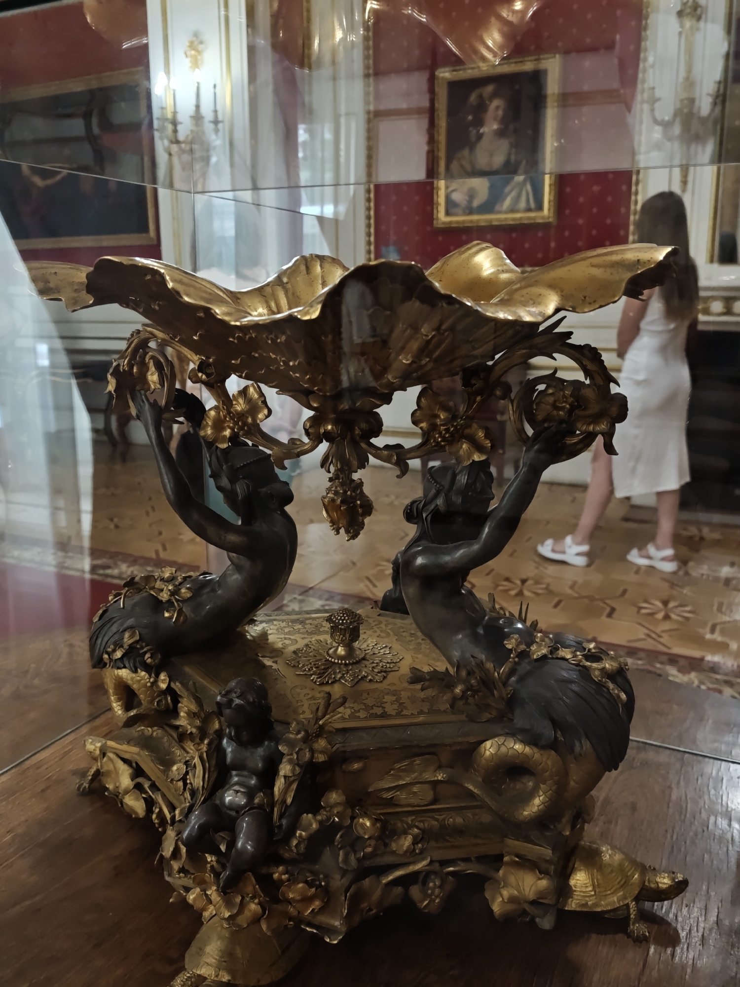 This is a medium shot, taken indoors, of a gilded ornamental centerpiece placed on a wooden table, likely within a museum or grand hall in Lviv, Ukraine. The centerpiece is highly ornate, featuring a large, shell-shaped bowl supported by two dark bronze figures of mermaid-like creatures. These figures have scaled tails and human torsos, and they are each holding decorative elements. The base of the centerpiece is also elaborate, adorned with floral motifs, a small cherubic figure, and what appears to be a turtle at the front. The object is displayed behind a glass barrier, which causes reflections of the surrounding room.
In the background, through the glass, a woman in a white dress and white sandals is visible, facing away from the camera and looking at an exhibition. Also visible are portions of framed artwork on red, patterned walls, and architectural details like ornate chandeliers and molding, suggesting a formal and historic setting. The flooring is a polished wood with inlaid patterns. The lighting appears to be a mix of ambient and possibly artificial light, creating highlights on the gilded parts of the centerpiece and contributing to the reflections in the glass.