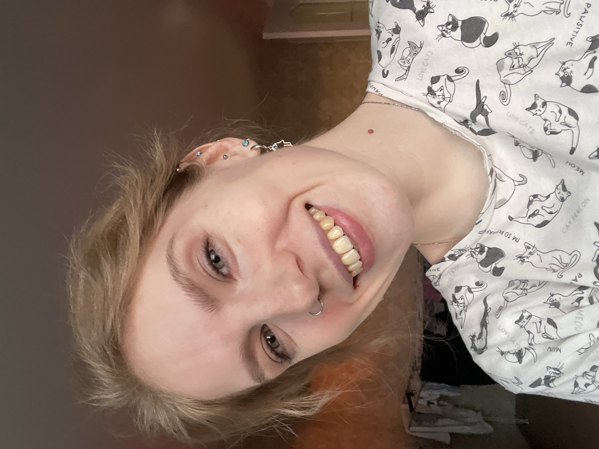 A close-up, canted portrait shows a smiling young woman with blonde hair. Her skin is fair, and she has light eyebrows and grey eyes. She has multiple ear piercings and a nose ring. Her teeth are slightly yellowed. She's wearing a white shirt with black and white cat drawings and printed text, including phrases like "PAWSITIVE," "CAT PERSON," "MEOW," "LOL CATS," and "I'M SO PURRFECT." The background is a blurry mix of grey and brown. There is no evidence to confirm this photo was taken in Izhevsk, Russia.