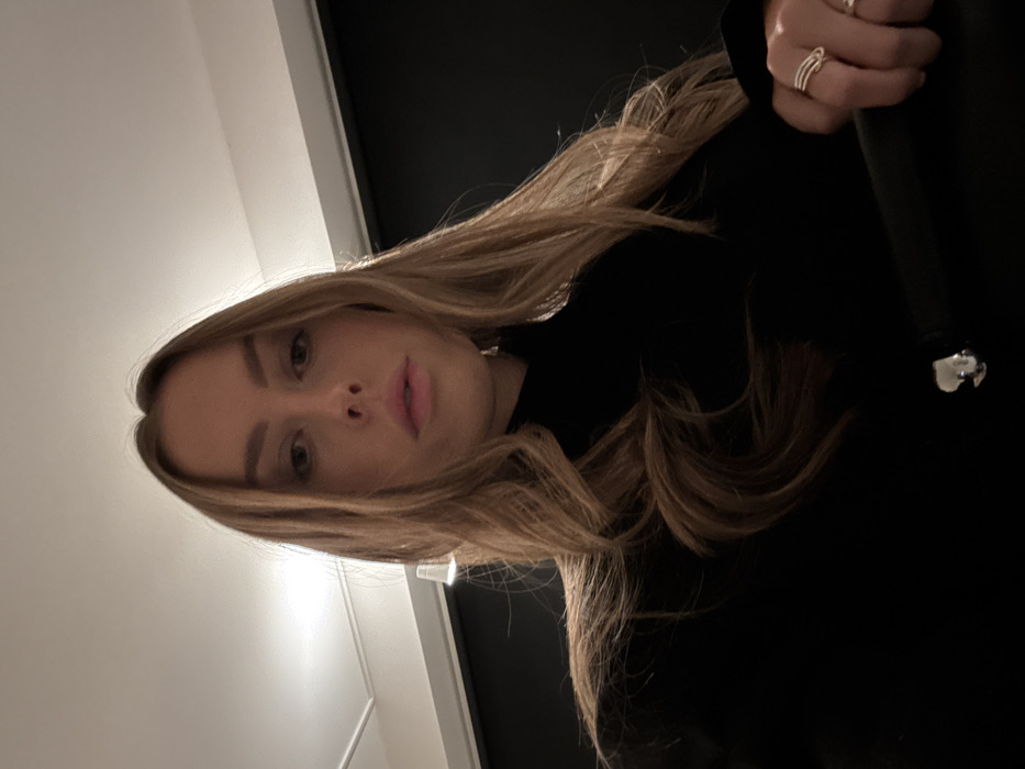 A fair-skinned woman with blonde hair framing her face gazes directly at the camera with a neutral expression. She is wearing a dark-colored top or dress, and her hand is visible, adorned with multiple rings. The background consists of a plain white wall contrasting with a dark wall, and possibly a doorframe. The photograph has a tilted perspective.