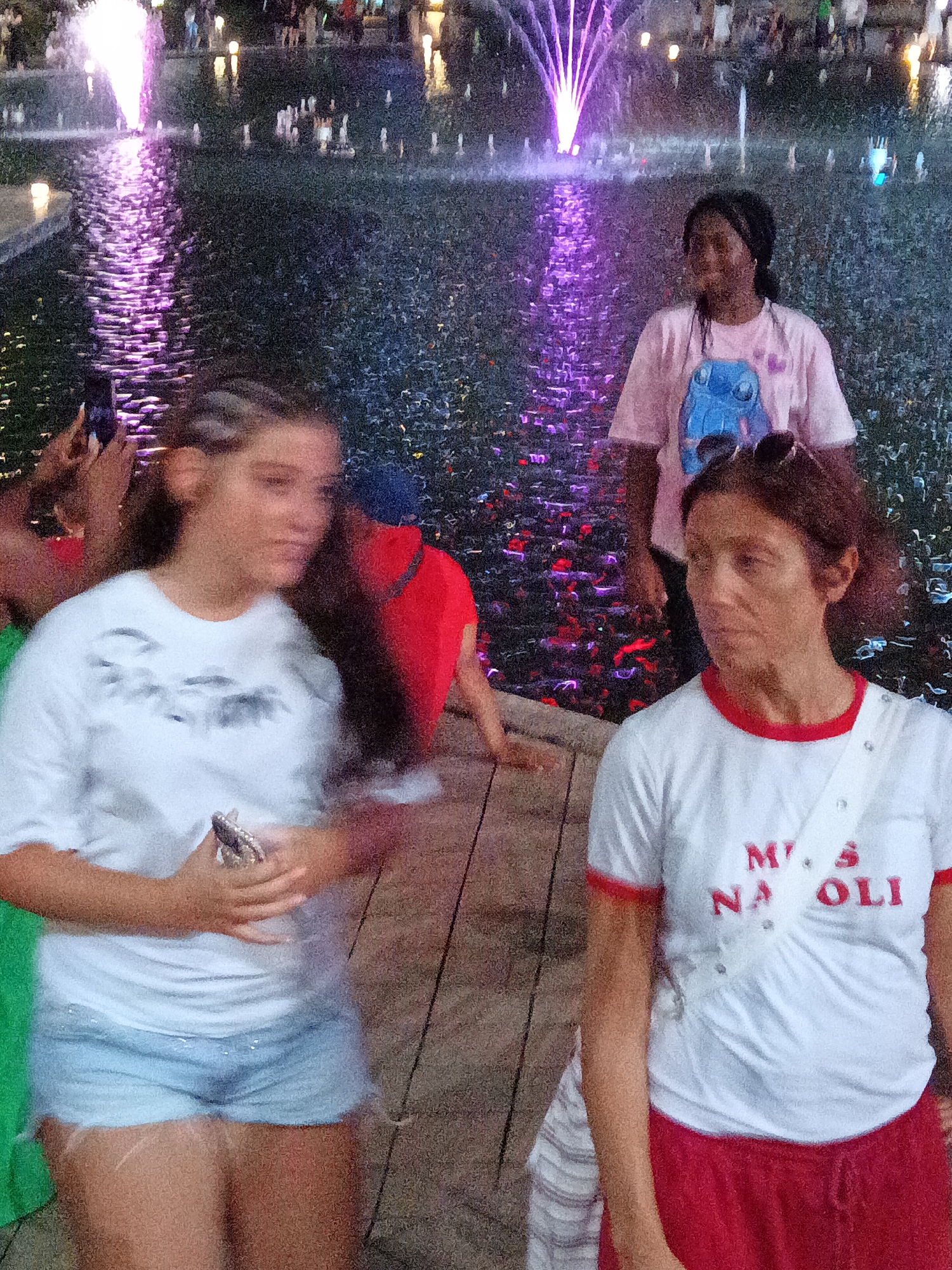 A group of individuals are near a fountain with purple lights at night. In the foreground, a young woman with braided hair is wearing a white t-shirt and light blue denim shorts, holding a phone. To her right, another woman in a white t-shirt with red trim and red shorts is looking to the side. Behind them, a person in a pink t-shirt with a cartoon character is visible. The background shows a water fountain with illuminated purple jets and other people observing. The scene appears to be an outdoor public space, possibly a park or entertainment area, in Kuala Lumpur, Malaysia.