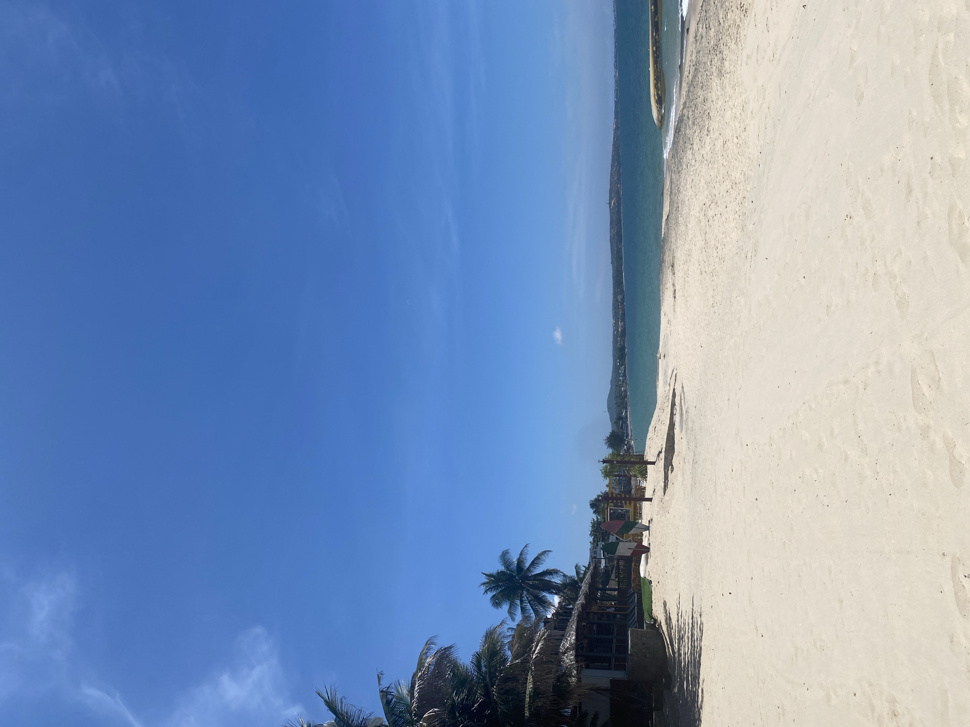 This is a photograph of a beach in Phu Thuy Ward, Vietnam. The scene is bathed in bright sunlight under a clear blue sky with scattered wispy clouds. The foreground is dominated by a wide expanse of light-colored sand, showing subtle footprints. To the left, a row of thatched-roof buildings, likely part of a resort or restaurant, are visible, shaded by palm trees. In front of these buildings, lounge chairs are arranged on the sand. Further into the beach, two surfboards, one red and white and the other green and white, stand upright. Beyond the beach, the turquoise ocean stretches to the horizon, meeting a distant coastline with hills and buildings. A curved breakwater or jetty is present in the water on the right side of the frame. The overall atmosphere is one of a tranquil tropical beach scene.