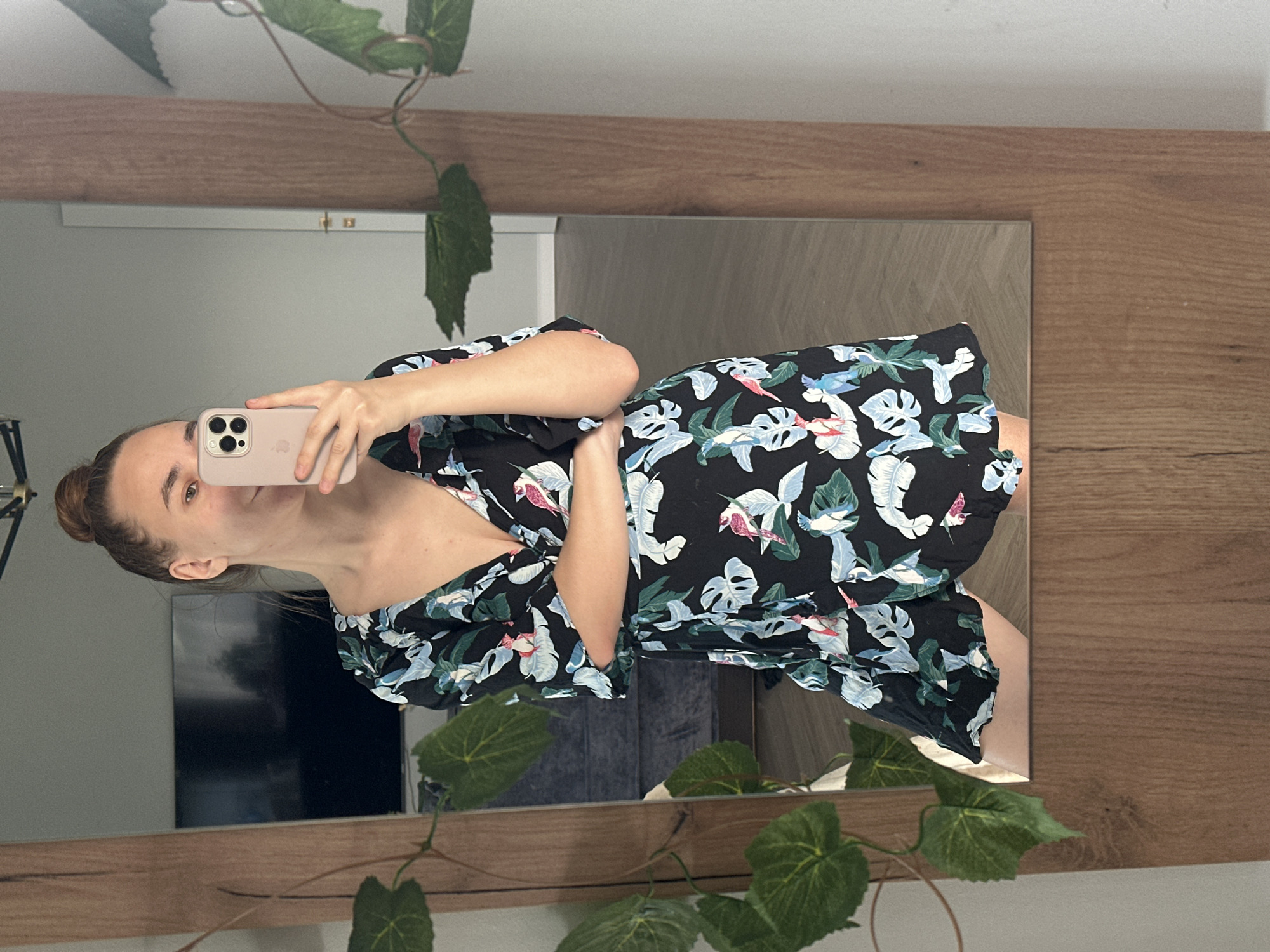 A woman with fair skin and brown hair in a bun stands in front of a mirror with a light-colored wooden frame. She is holding a pink smartphone in her left hand, presumably taking a selfie. She is wearing a black dress with a floral pattern featuring white, green, and blue leaves and pink birds. The mirror reflects a living room setting, including a television mounted on the wall and a grey sofa. A potted plant with green leaves partially obscures the mirror’s reflection. The location is not explicitly stated but is assumed to be Kharnavy, Russia.