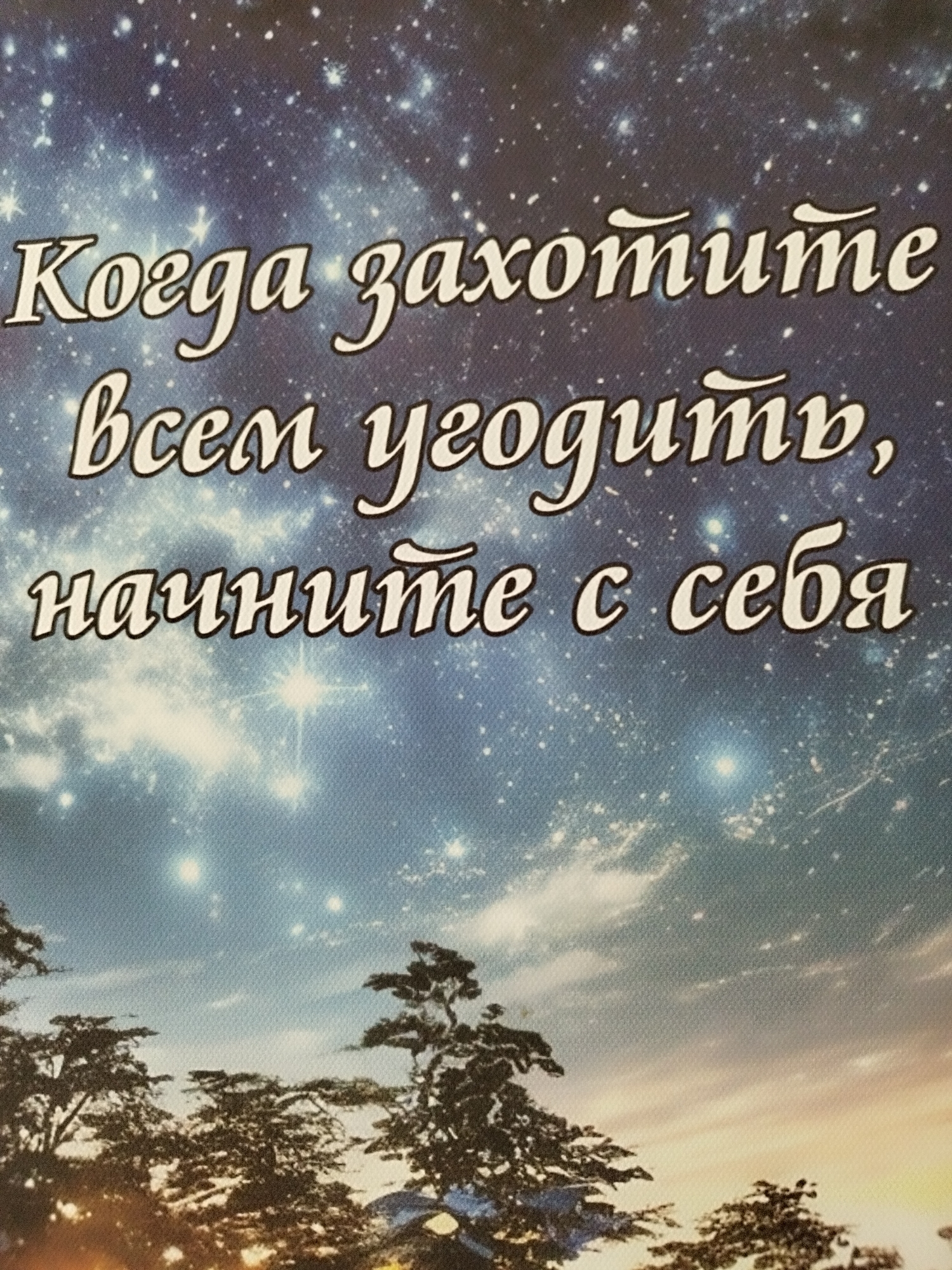 This image shows a celestial scene with trees silhouetted against a starry sky. The text on the image reads "Когда захотите всем угодить, начните с себя," which translates to "When you want to please everyone, start with yourself." The sky is a deep blue, speckled with numerous stars and some wispy clouds, suggesting nighttime. The trees are dark against the lighter sky, with some branches reaching upwards. The overall impression is one of contemplation and self-reflection, possibly linked to the inspirational quote. There are no people or specific objects visible beyond the trees and the text. The lighting and colors suggest a serene and possibly a touch of melancholy atmosphere. The location context provided is Skadovsk, Ukraine, though there are no explicit visual cues in the image to confirm this specific location.