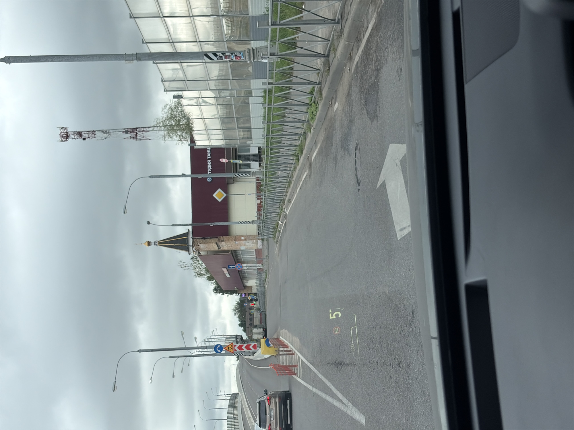 The image shows a road view from inside a vehicle in Balashikha, Russia. The road is asphalt with white lane markings. On the right side of the road, there is a metal fence, followed by a transparent sound barrier. A telecommunications tower is visible in the distance. On the left side of the road, there is a curved overpass and a dark colored SUV. A building with a maroon roof and text that appears to read "СТУДИЯ ТАНЦ" (Dance Studio) is visible. There are streetlights lining the road. A speed limit sign of 60 is visible, along with a digital display showing a speed of 57. The sky is overcast with clouds.