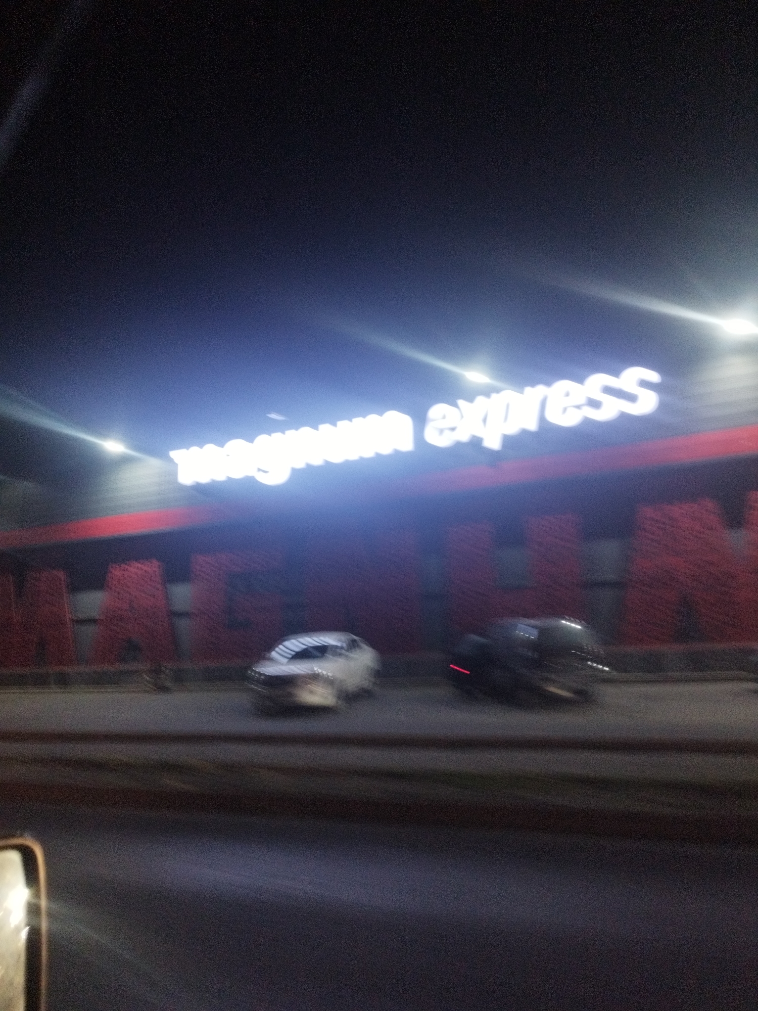 The image shows a nighttime view of a building facade with a prominent neon sign that reads "magnum express." Below the sign, large red letters are visible, which appear to spell out "MAGNUM" though they are somewhat blurred. Two cars are on the road in front of the building; one is a white sedan, and the other is a dark-colored SUV or van. The image is taken from a moving vehicle, as indicated by the blurriness and the partial view of a side-view mirror in the lower left corner. The environment appears to be an urban setting, likely a commercial area, given the signage and the presence of vehicles. The sky is dark, suggesting it is nighttime.