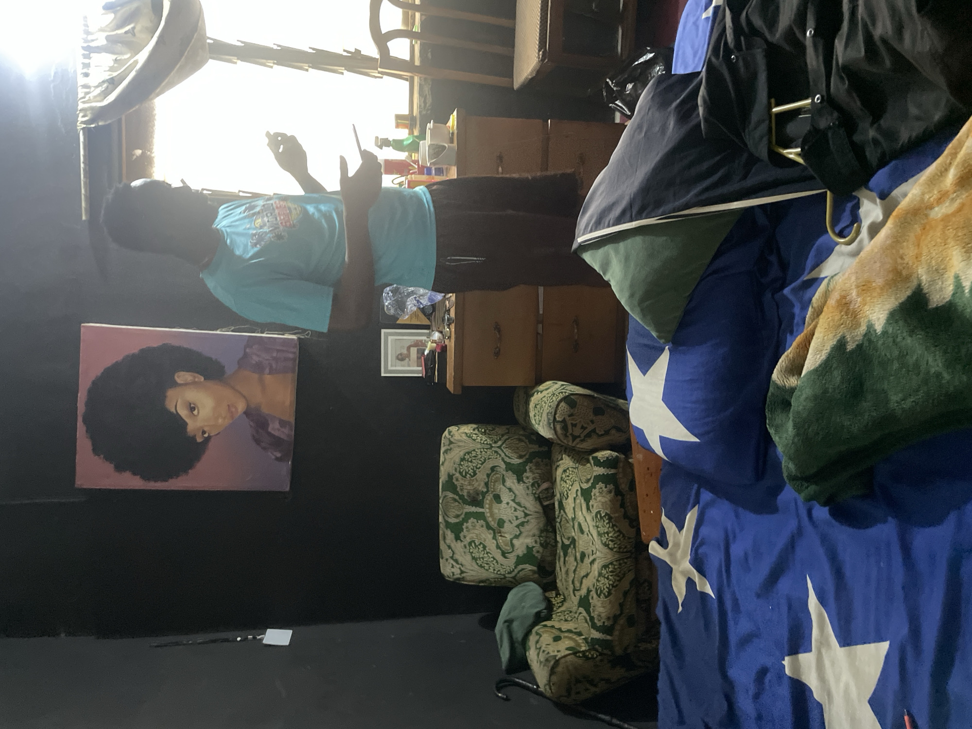 The image depicts an interior scene, likely a bedroom, in Jos, Nigeria. In the background, a portrait of a woman with an afro hangs on a black wall. To the right, a man in a light blue t-shirt and dark shorts stands near a window. He is holding a mobile device. A wooden dresser with drawers is positioned below the window, with various items on its surface including picture frames, bottles, and a bucket. Adjacent to the dresser is a wooden chair. In the foreground, a floral-patterned armchair is visible. A bed is spread with a blue blanket featuring white star patterns. Pillows, one green and one dark blue, are on the bed, along with a folded blanket in shades of green and orange. A black jacket is draped over a clothes hanger on the bed. The lighting suggests it is daytime, with sunlight streaming through the window.