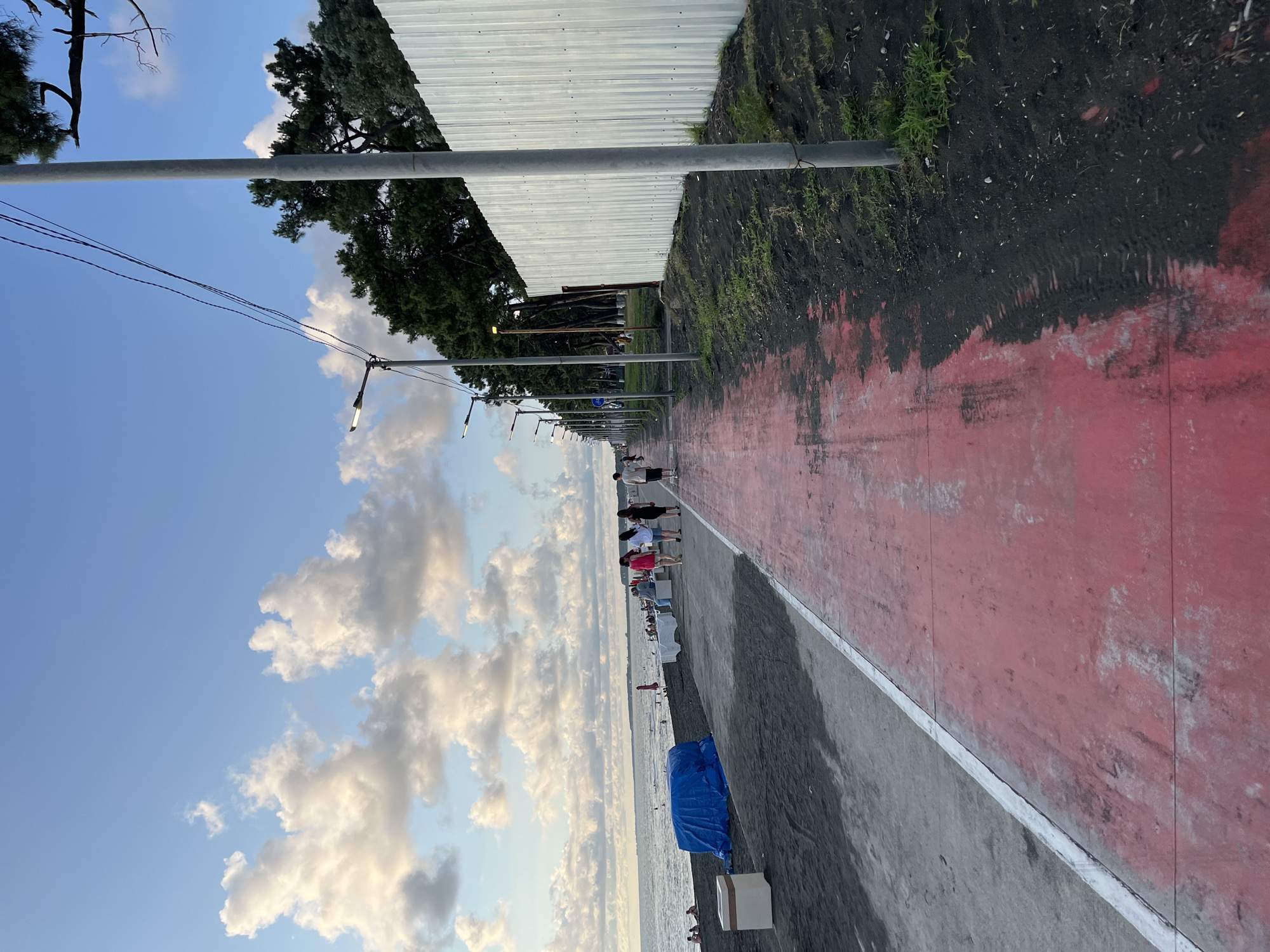 The image depicts a seaside promenade in Ureki, Georgia, on a partly cloudy day. The promenade is paved with a red surface for walking or cycling, with a gray strip bordering the beach. To the left, a dark sand beach meets the calm sea, with several people visible in the distance, some swimming or sunbathing. A large object covered in a blue tarp is situated on the beach. Along the right side of the promenade, a line of tall trees stands behind a corrugated metal fence. Several streetlights and power lines are visible along the promenade. A few individuals are walking along the promenade. The sky is a vibrant blue with scattered white clouds, suggesting favorable weather.