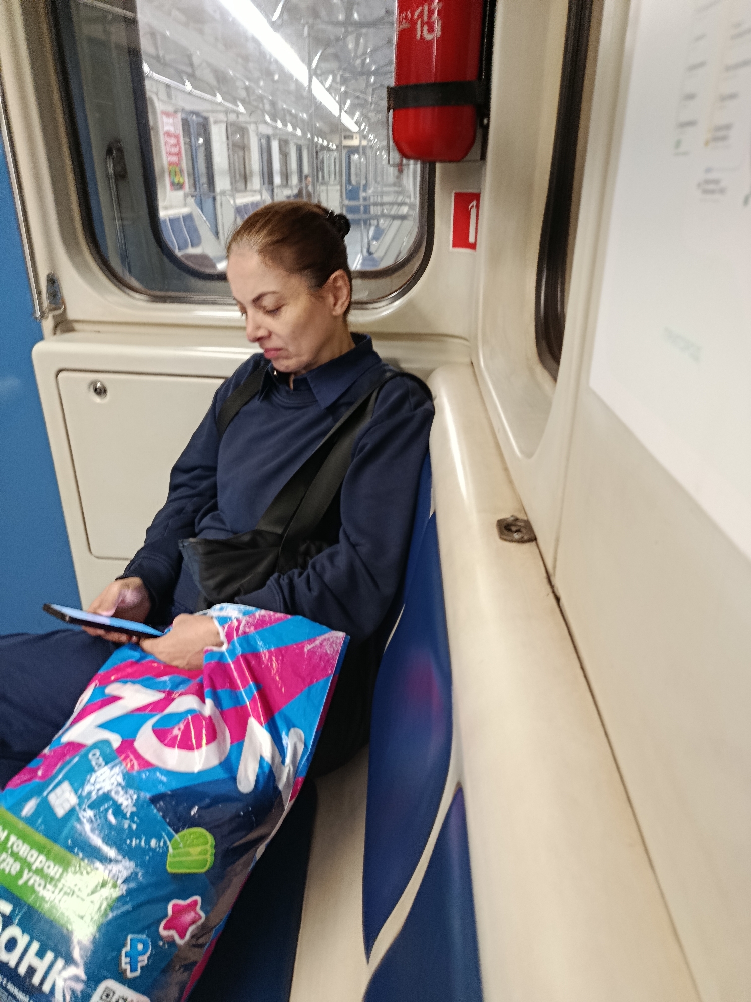 A woman with brown hair pulled back in a bun is seated on a blue and white upholstered bench inside a Moscow Metro train car, looking down at her smartphone. She is wearing a dark blue long-sleeved shirt and dark blue pants, with a black bag slung across her chest. She holds a brightly colored shopping bag, predominantly pink and blue with white lettering, in her lap. The train car interior is beige and blue, with windows showing the interior of another train car passing by. A red fire extinguisher is mounted on the wall above and to the right of the woman, and a red square with a white numeral "1" is visible to its left. The scene is illuminated by overhead lighting.
