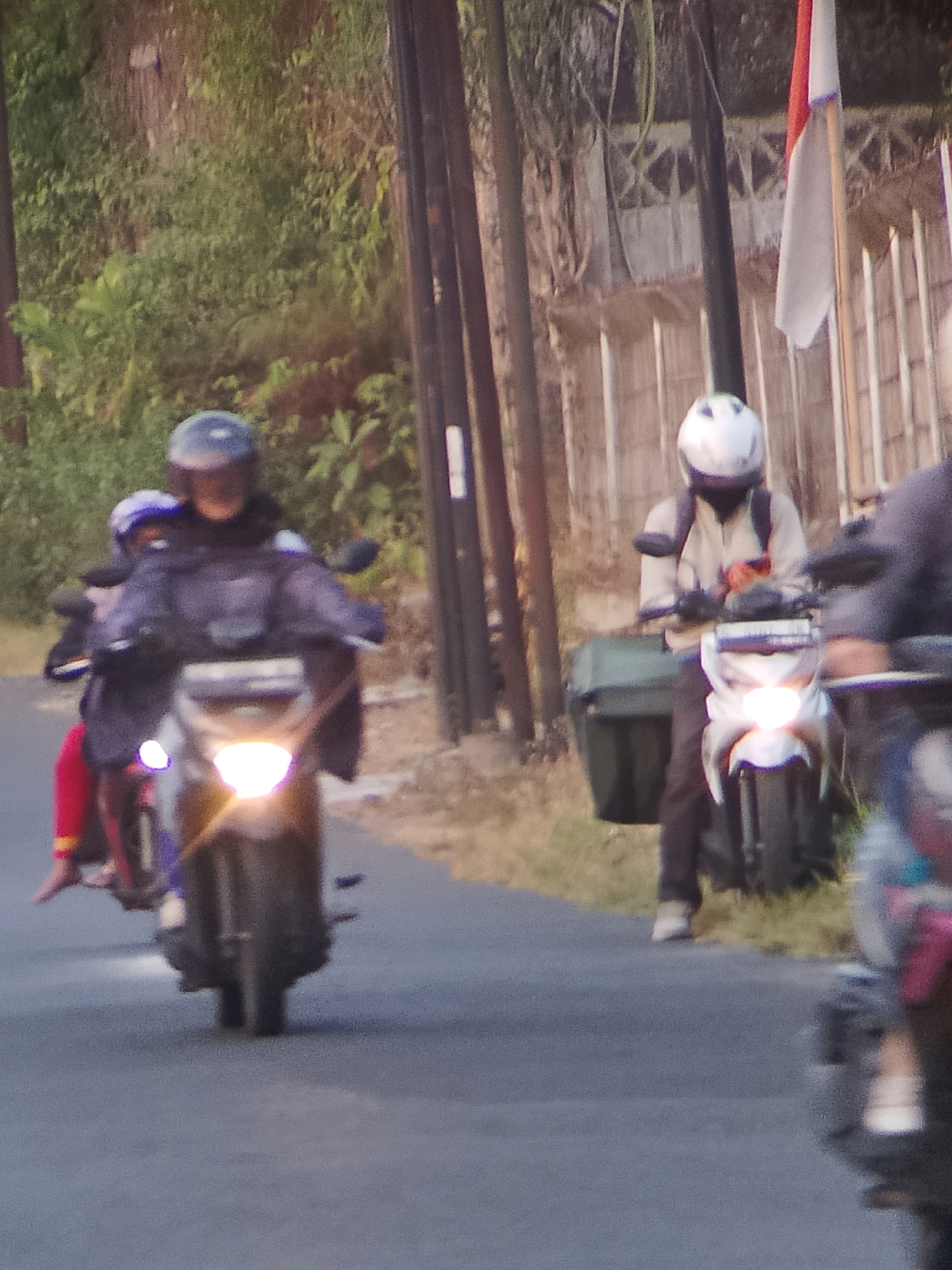 This image captures a street scene in Jember, Indonesia, featuring several individuals on motorcycles. In the foreground, two people ride a dark-colored motorcycle, with the rider wearing a dark helmet and jacket. The passenger behind them has a purple helmet and a red garment. The motorcycle's headlight is illuminated. Further down the road, another motorcycle is visible, ridden by a person wearing a white helmet and a light-colored jacket, with a dark backpack. A dark green storage box is attached to this motorcycle. The background consists of vegetation and a fence, with a pole and a flag visible. The road surface is asphalt.