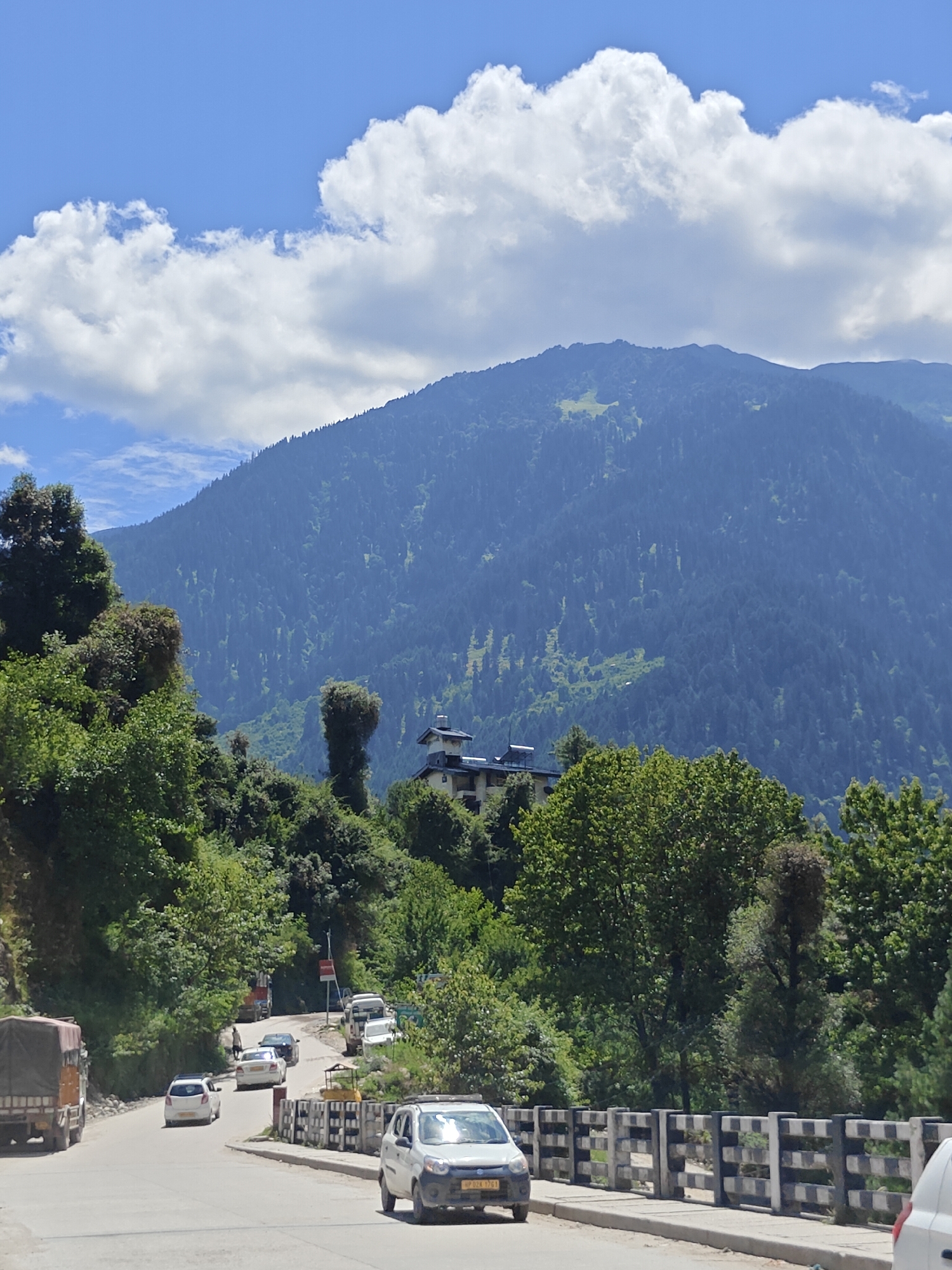 This is a photograph of a road in Aleo, India, with a mountain backdrop. The road curves to the left, with several vehicles on it. A white car is in the foreground, and a truck with a canvas cover is on the left side of the road. Further up the road, there are more cars, including a white hatchback and a dark sedan. A single pedestrian is walking up the road, in front of the dark sedan. The road is lined with trees on the left, and a hillside covered in dense evergreen forest is visible on the right. A building complex with a white facade and dark roofs is situated on the hillside. The sky is blue with fluffy white clouds. The overall scene depicts a typical mountain road scene in a populated area.