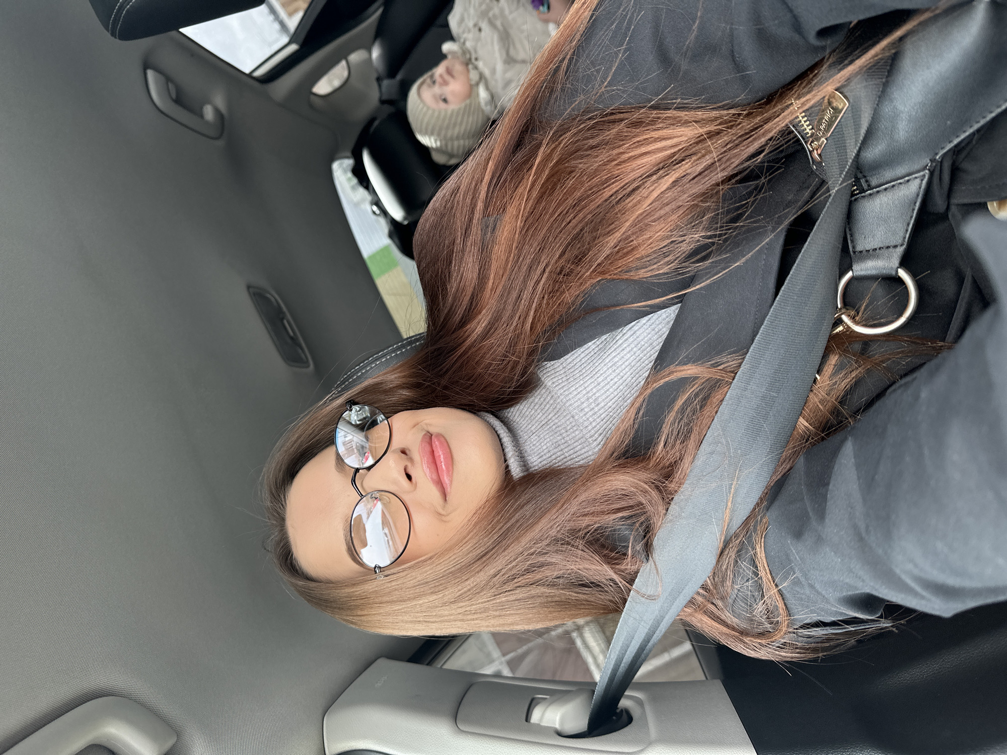 The image, oriented as if viewed upright, captures a young woman seated in the passenger side of a car, looking directly at the camera with a slight smile. She has long, straight reddish-brown hair that cascades over her shoulders and chest. She is wearing round, wire-rimmed glasses, a light gray turtleneck sweater, and a dark, possibly black, jacket. A gray car seatbelt diagonally crosses her chest and lap.
Behind her, in the back seat, a young child or infant is visible in a car seat, wearing a light beige or off-white knit hat that covers their head and ears. Only the top portion of the child's face and head are clearly discernible.
The car's interior features a light gray headliner with a grab handle visible towards the upper left. A sunroof or panoramic roof is partially visible, indicating an open or bright overhead view. Through the car windows, a muted outdoor scene suggests an overcast sky or winter conditions, aligning with the location context of Yekaterinburg, Russia.
A black bag strap with a metallic zipper is visible over the woman's right shoulder, with a round silver-colored metal ring attached. Indiscernible text is partially visible on the zipper pull, which, upon closer inspection in the crops, appears to read "BURBITGI". The overall scene depicts a casual daytime moment inside a vehicle.