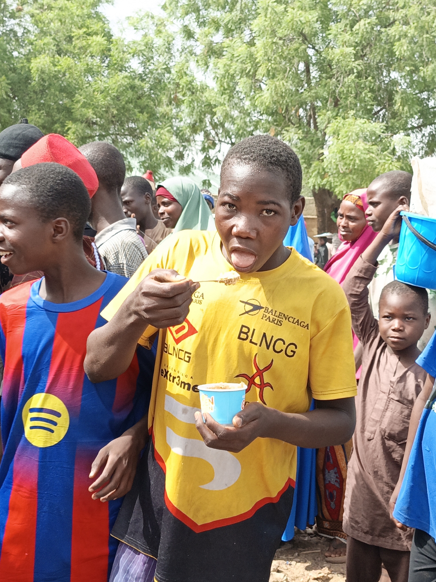 In an outdoor setting in what is suspected to be Kawaji, Nigeria, a crowd of people stand beneath a tree. A young person in a yellow Balenciaga t-shirt licks food from a spoon while holding a blue cup. To their left, another youth wears a red and blue Spotify t-shirt. Behind them are several individuals, including one in a green headscarf and another in a pink headscarf. A child wearing a brown garment stands holding a blue bucket. The ground is dirt.