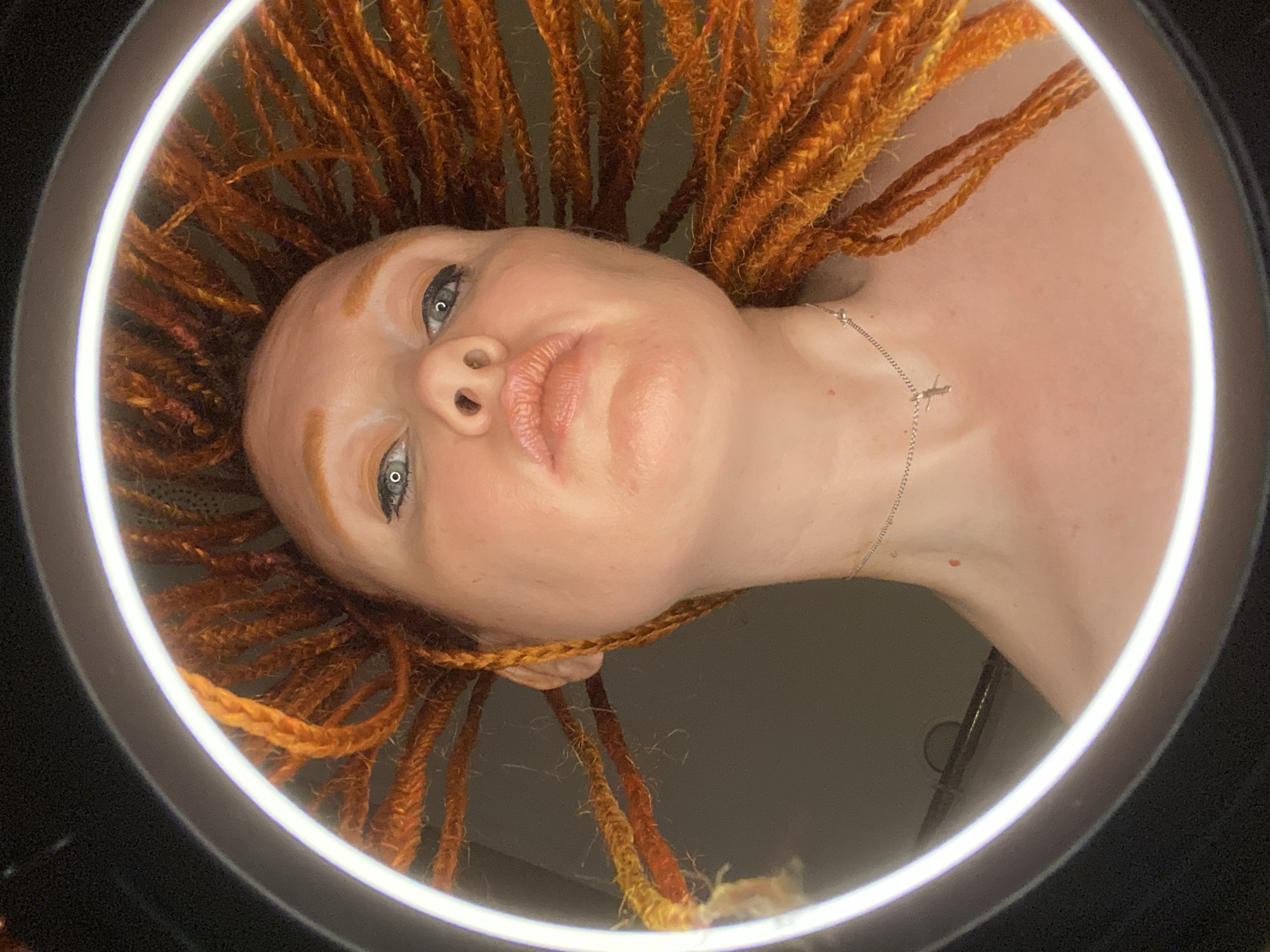 A low-angle, close-up shot shows a woman from the chest up, viewed through a ring light. The woman has bright orange braided hair that fans out around her head. She is wearing a silver necklace with a small pendant. Her eyes are open and looking upwards, and her lips are slightly pursed. The lighting is bright, creating a halo effect around her head. The background is dark and out of focus. There is no information in the image to suggest a location in the city of Sochi, Russia.