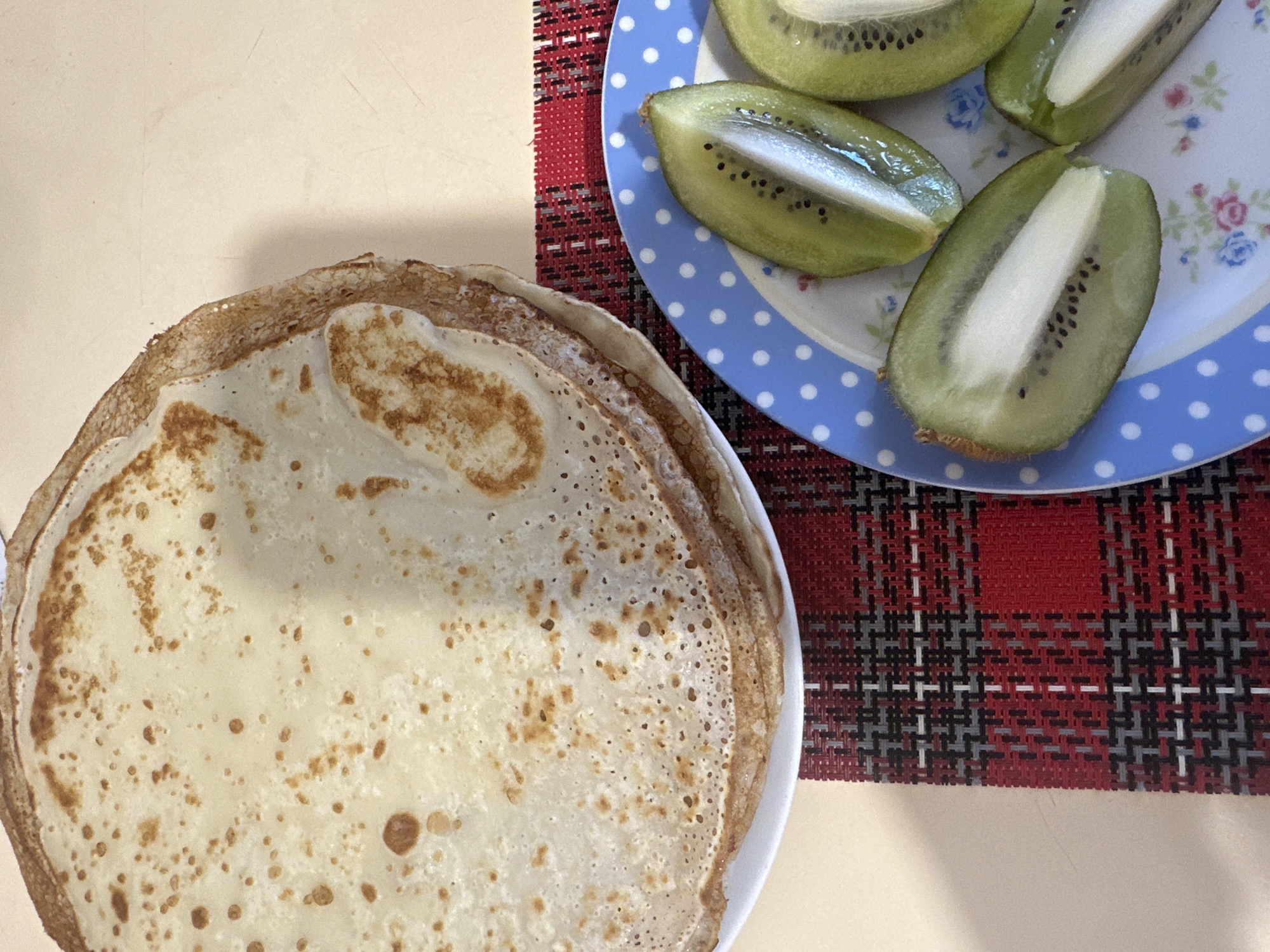 A top-down shot shows a stack of pancakes on a white plate and slices of kiwi fruit on a blue and white polka-dot plate. The scene is set on a surface with a patterned placemat, displaying red, black, gray, and white interwoven lines. The background is a pale beige or cream color. No people are visible. This image does not provide enough information to discern the location in the city of Votkinsk, Russia.