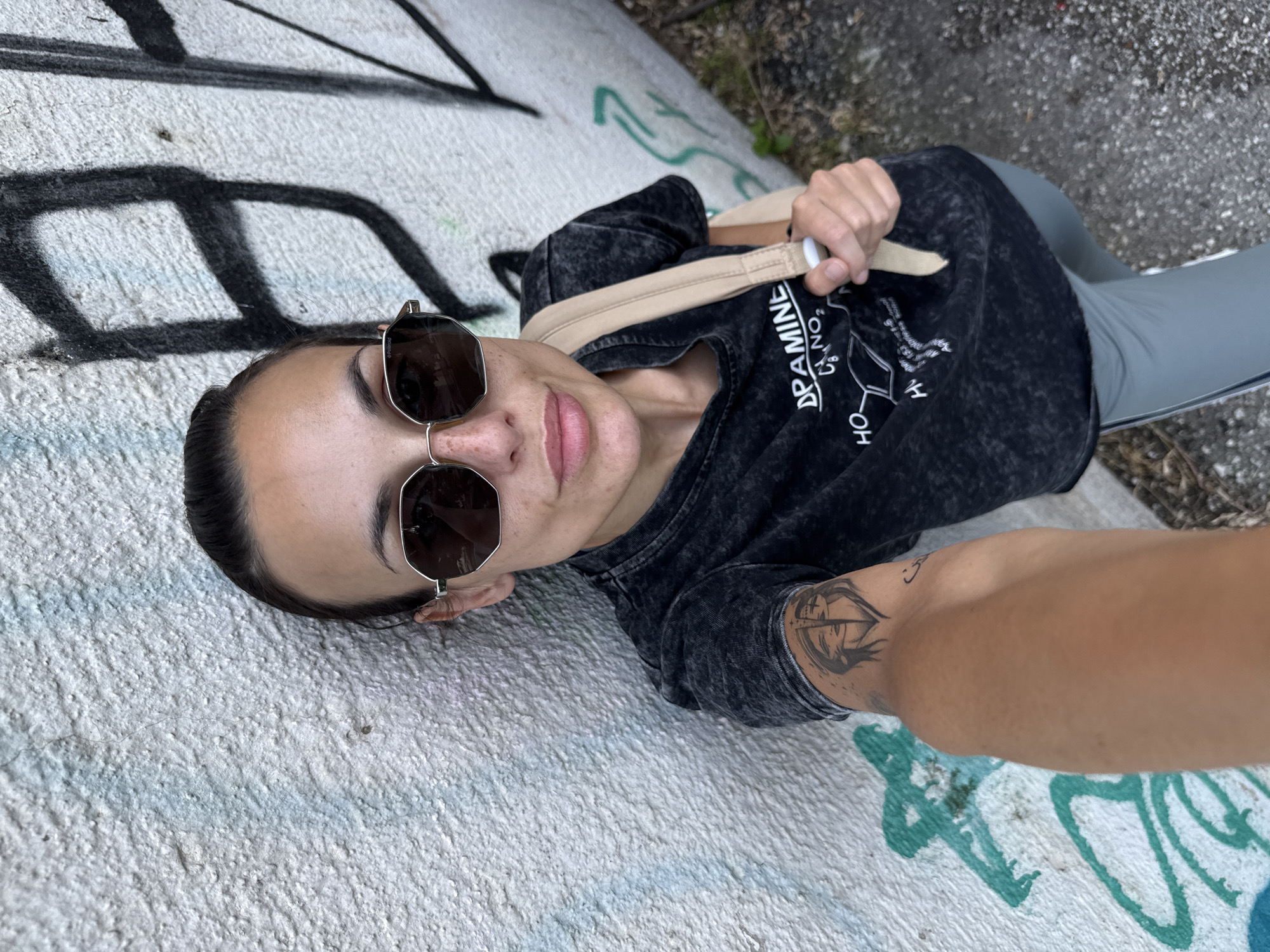 A person, likely female, is lying on a concrete surface covered in graffiti. She is wearing sunglasses, a dark T-shirt with a chemical formula printed on it (likely dopamine), and grey leggings. She is holding the strap of a beige bag. There is a tattoo visible on her right arm. The location is inferred to be in Pleven, Bulgaria, based on the request. The scene appears to be a casual selfie taken outdoors.