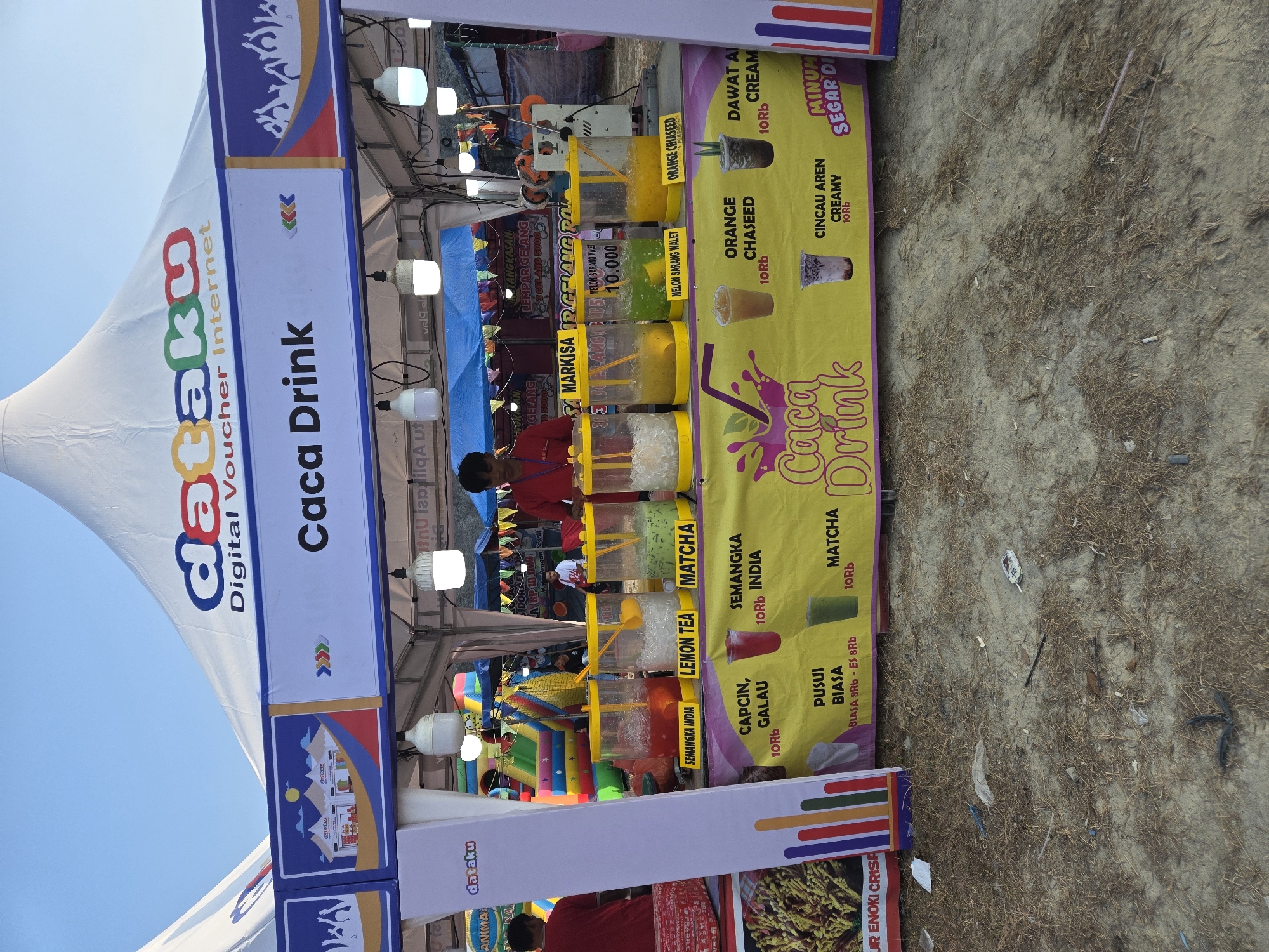 A roadside stall in Sungailiat, Indonesia, is set up under a white tent with "Dataku Digital Voucher Internet" branding. The stall, identified as "Caca Drink," displays various drinks in clear plastic containers filled with ice and colorful liquids. Labeled options include Semangka India, Lemon Tea, Matcha, Orange Chased, and Cincau Aren Creamy. Menu boards indicate prices in Indonesian Rupiah, with most drinks priced at 10Rb. A vendor wearing a red shirt stands behind the counter, serving customers. In the background, an inflatable playground can be seen. The ground is covered with dry grass and sand, suggesting an outdoor, possibly beachside, location. Trash is scattered on the ground.