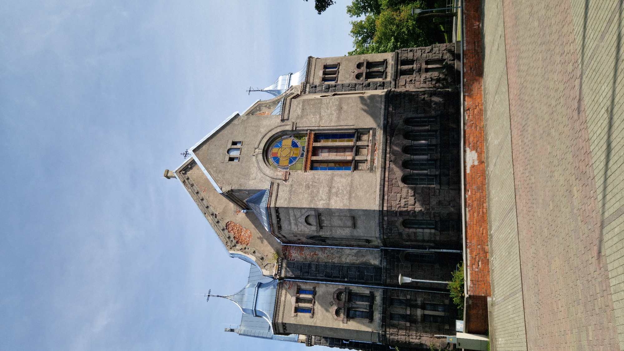The image shows a large, stone building with a distinctive blue-tinted metal roof, possibly a church or public building, located in Liepaja, Latvia. The building features a large, arched stained-glass window with a circular design and several rectangular stained-glass windows below it. The stonework of the building appears weathered, with some areas showing exposed brick and damage. In the foreground, there is a paved area with a brick border and a lamp post. The sky is clear with a few faint clouds.