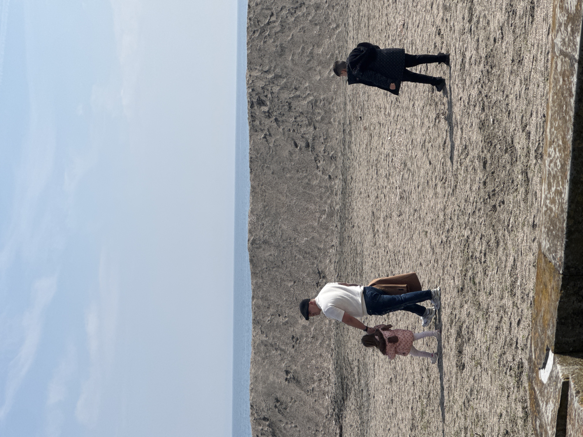 The image shows a coastal scene, likely a man-made structure on the coast of Burgas, Bulgaria. The left side features the sky and the sea. On the right, there are two adults and a child. One adult is walking alone in a dark jacket, while another adult is walking with a child. The adult and child are further away from the edge. They're walking on a rocky, tan, textured surface. A partial stone structure with moss or white marks is partially visible on the right side of the image.