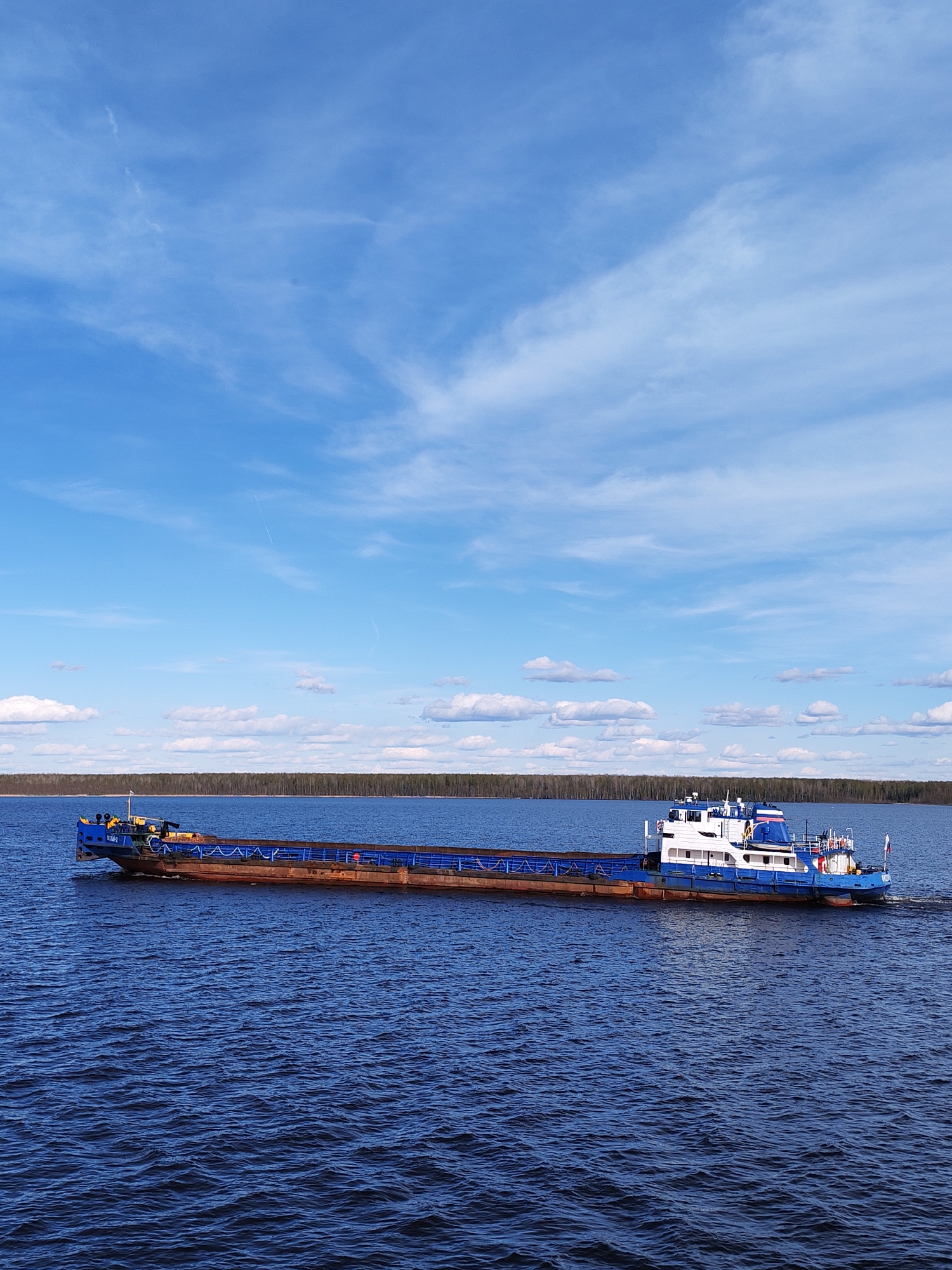 A long blue and brown barge moves across a body of water under a partly cloudy blue sky. The barge has a blue tugboat section at the front and the back. The body of water extends to the horizon where there is a thin line of trees. The location is Gorushka, Russia.