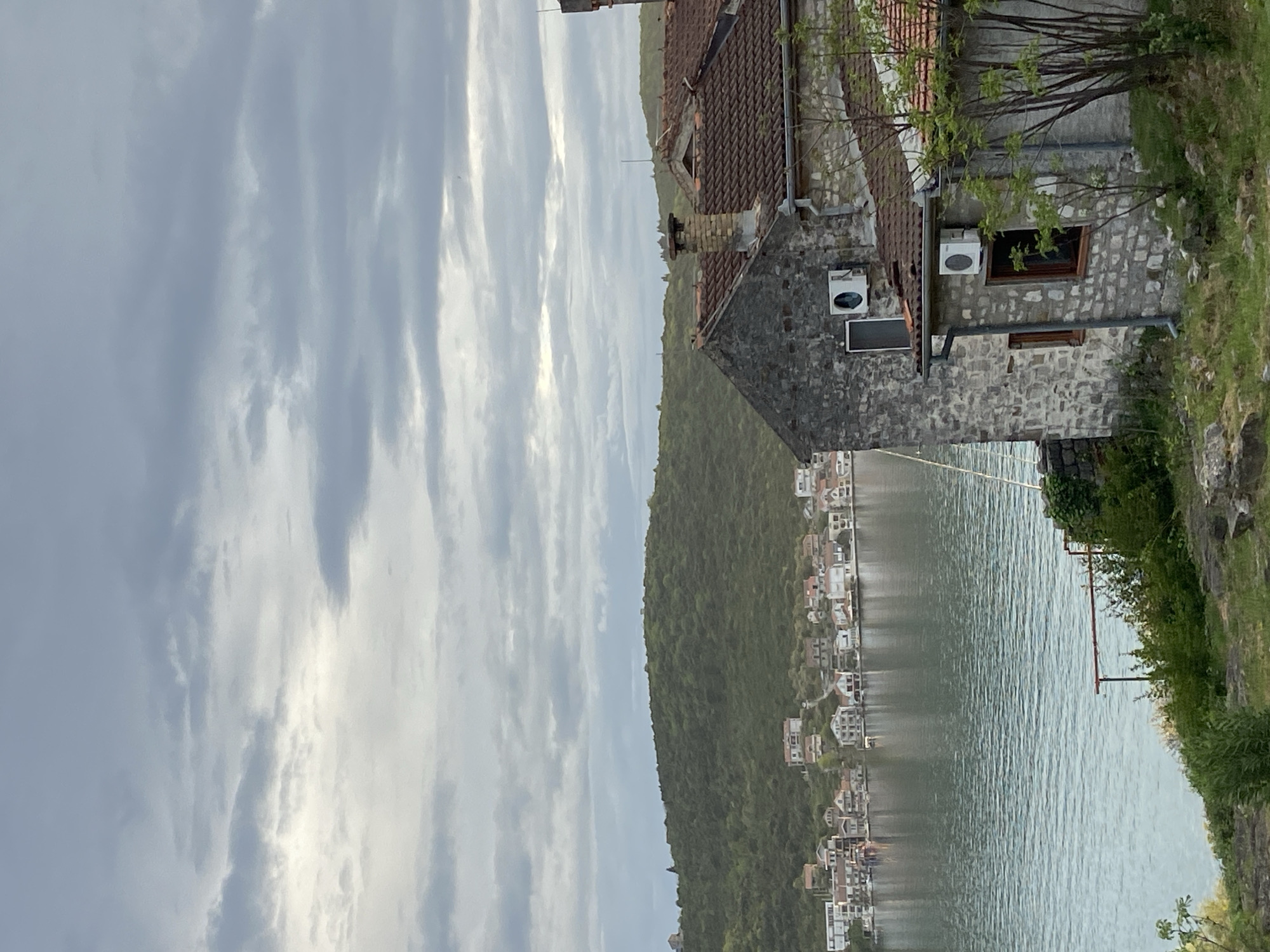 The image depicts a waterfront view in Đuraševići, Montenegro. In the foreground stands a stone building with a brown tiled roof, featuring air conditioning units and windows. Adjacent to the building is a grassy area. The middle ground shows a body of water with the reflection of houses lining the opposite shore, backed by a forested hillside. The sky is overcast with elongated cloud formations. The scene is still and appears to be daytime.