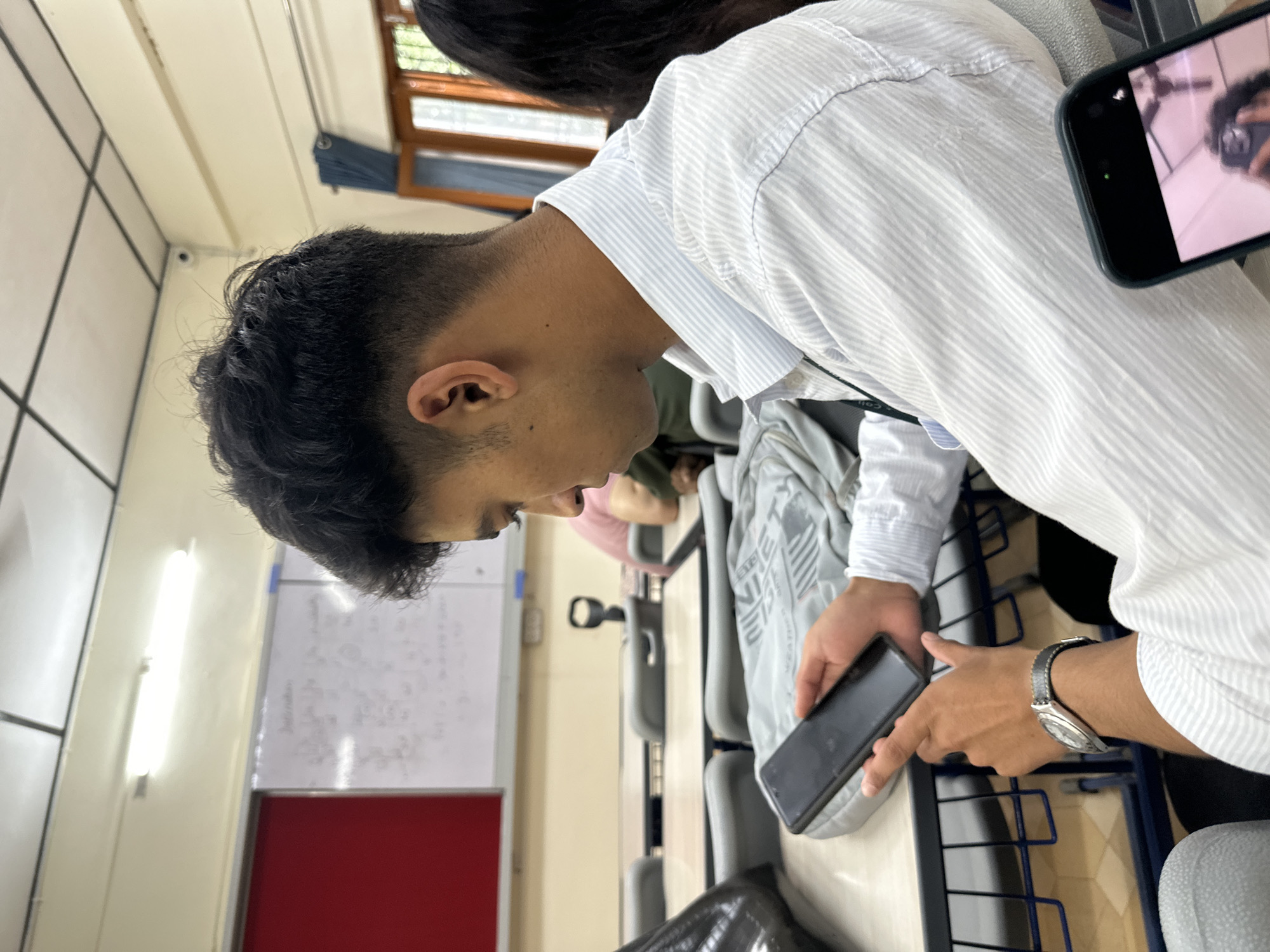 A young man is seated at a desk, looking down at his smartphone. He is wearing a white striped shirt and a watch. A bag is on the desk next to him. In the background, a whiteboard is visible, along with other students seated at desks. The environment appears to be a classroom or lecture hall. The room is lit by fluorescent lights and has windows on one side.