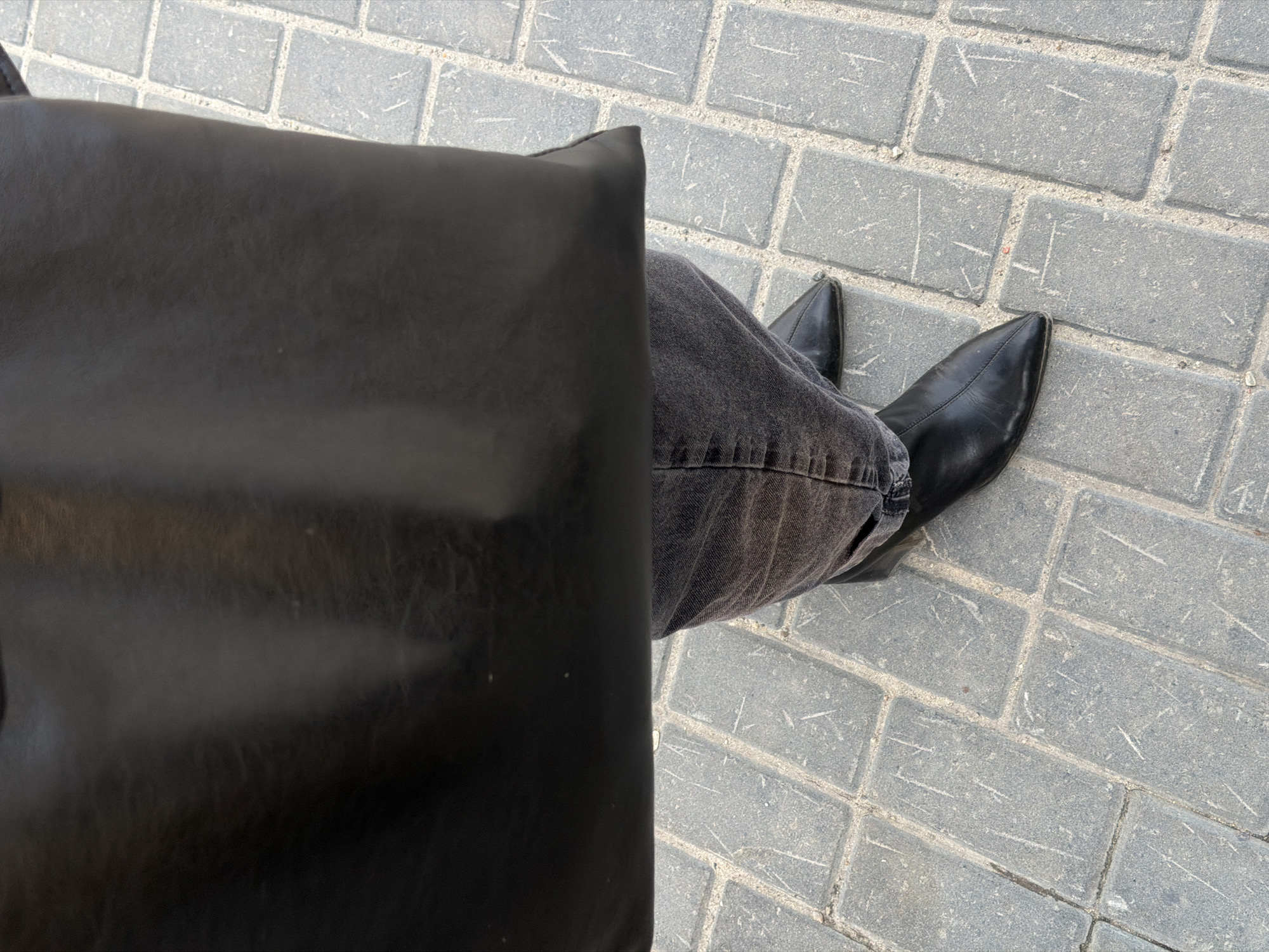 A top-down shot shows a person seated outdoors in Засечное, Russia. The ground is paved with grey rectangular bricks. The person is wearing grey jeans and black leather pointed-toe boots. A large black leather bag sits next to the person, obscuring part of their legs.