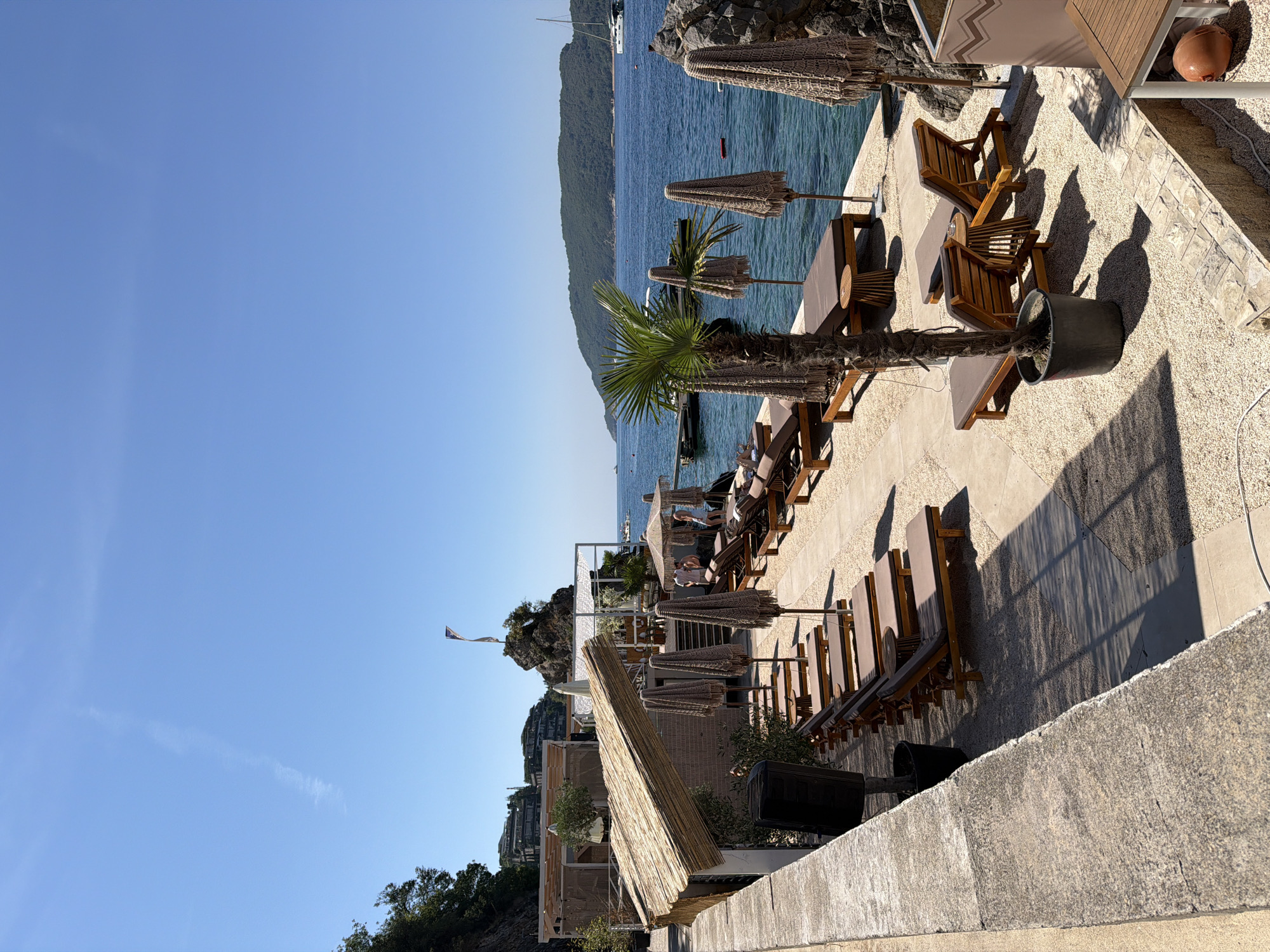 The image depicts a view of a beach club with lounge chairs and umbrellas arranged on a gravelly area adjacent to a calm, blue sea. In the background, a lush green hillside with buildings is visible, and a sailboat is anchored in the distance. The sky is clear and bright blue. The scene appears to be located in Budva, Montenegro, likely at a popular seaside resort. The overall atmosphere is relaxed and inviting, suggesting a sunny day at the beach.