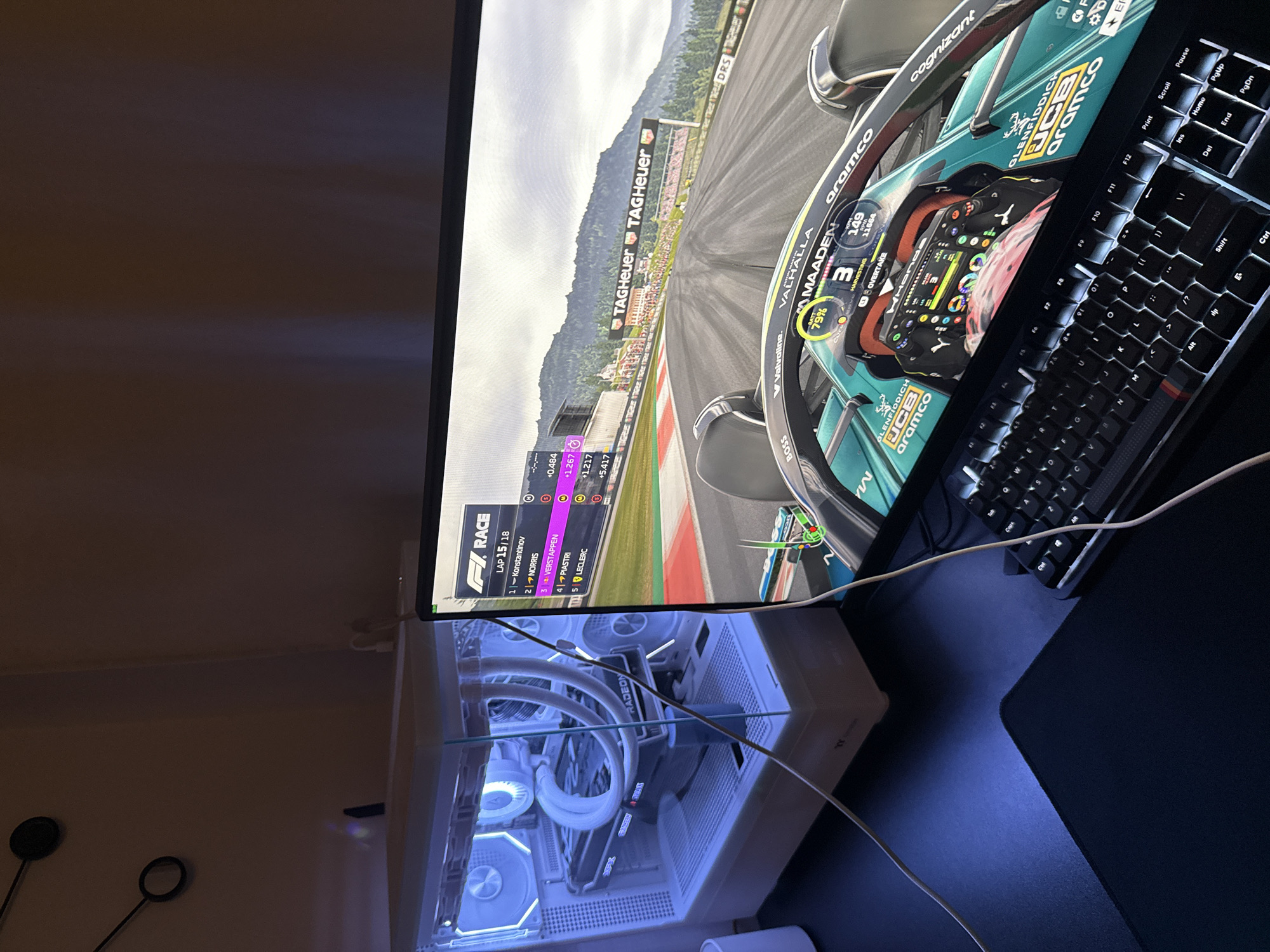 Here is a forensic description of the image:
The image shows a computer setup featuring a monitor displaying a Formula 1 racing game, a white computer tower with visible internal components illuminated by blue LED lighting, and a black mechanical keyboard placed on a dark surface, possibly a desk with a black mouse pad. The game is in progress with a race leaderboard visible on the left side of the monitor. The car displayed on the monitor is adorned with logos from "aramco" and "cognizant" among other sponsorships. The leaderboard indicates that the current lap is 15 of 18. The monitor appears to be positioned on top of the computer tower, with cables extending towards the keyboard and the surrounding area. The scene is set indoors, potentially in a room within a residence. The location in the city of Unknown, Unknown cannot be determined from the information provided.