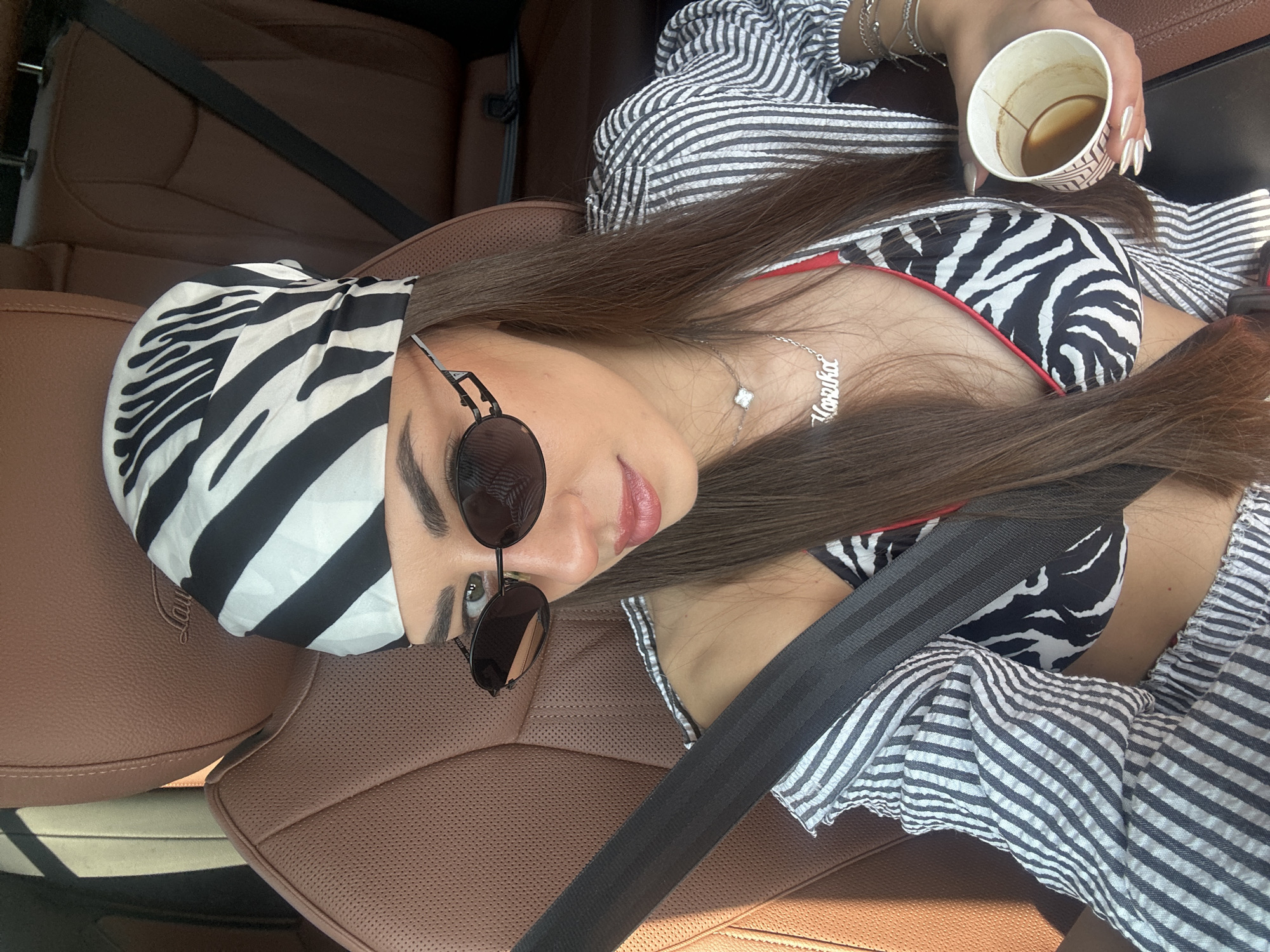 A woman with dark hair, wearing a zebra-print bandana and sunglasses, is seated in the driver's seat of a car. She is wearing a zebra-print bikini top and a striped button-up shirt, with a silver necklace and bracelets. She holds a paper cup of coffee in her right hand. The car interior appears to be leather, with a brown headrest visible. The scene is lit by sunlight streaming in from the left. The location is specified as Neo Souli, Greece.