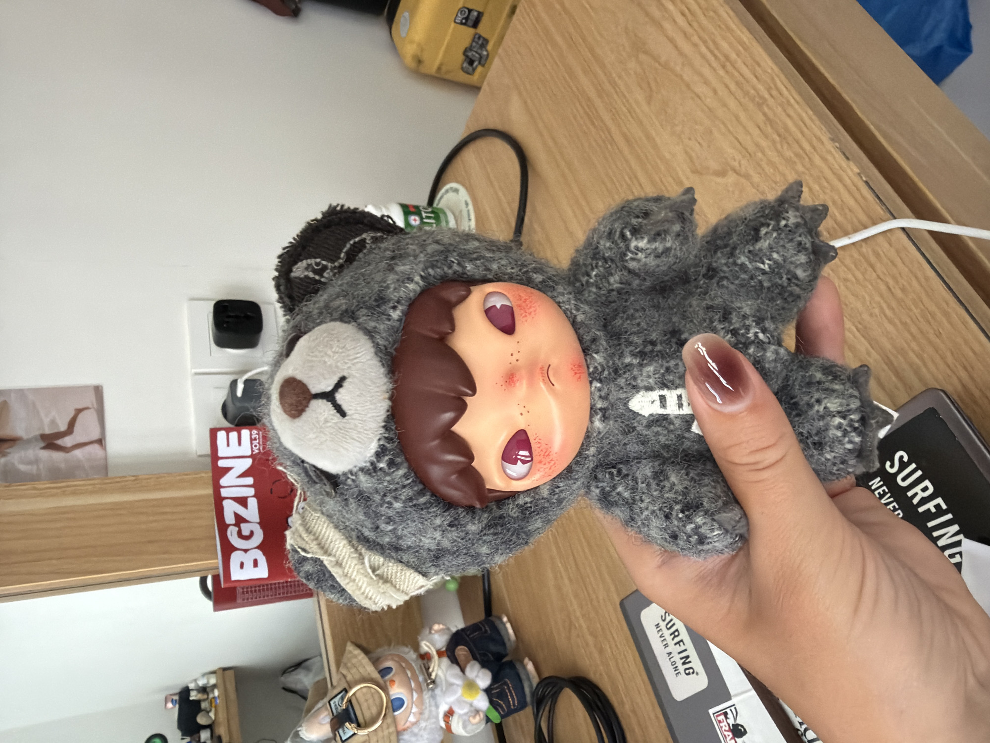 A human hand holds a gray stuffed animal, featuring a bear's head with a human-like face, against a light brown wooden surface. The face has brown hair and a peachy complexion, with red markings on the cheeks. The bear head is primarily white, with a gray textured body. Adjacent to the hand and stuffed animal are a gray and black book titled "SURFING NEVER ALONE", a red box with "BGZINE" printed on it, and various small objects. The location is indicated to be Kuta Selatan, Indonesia.