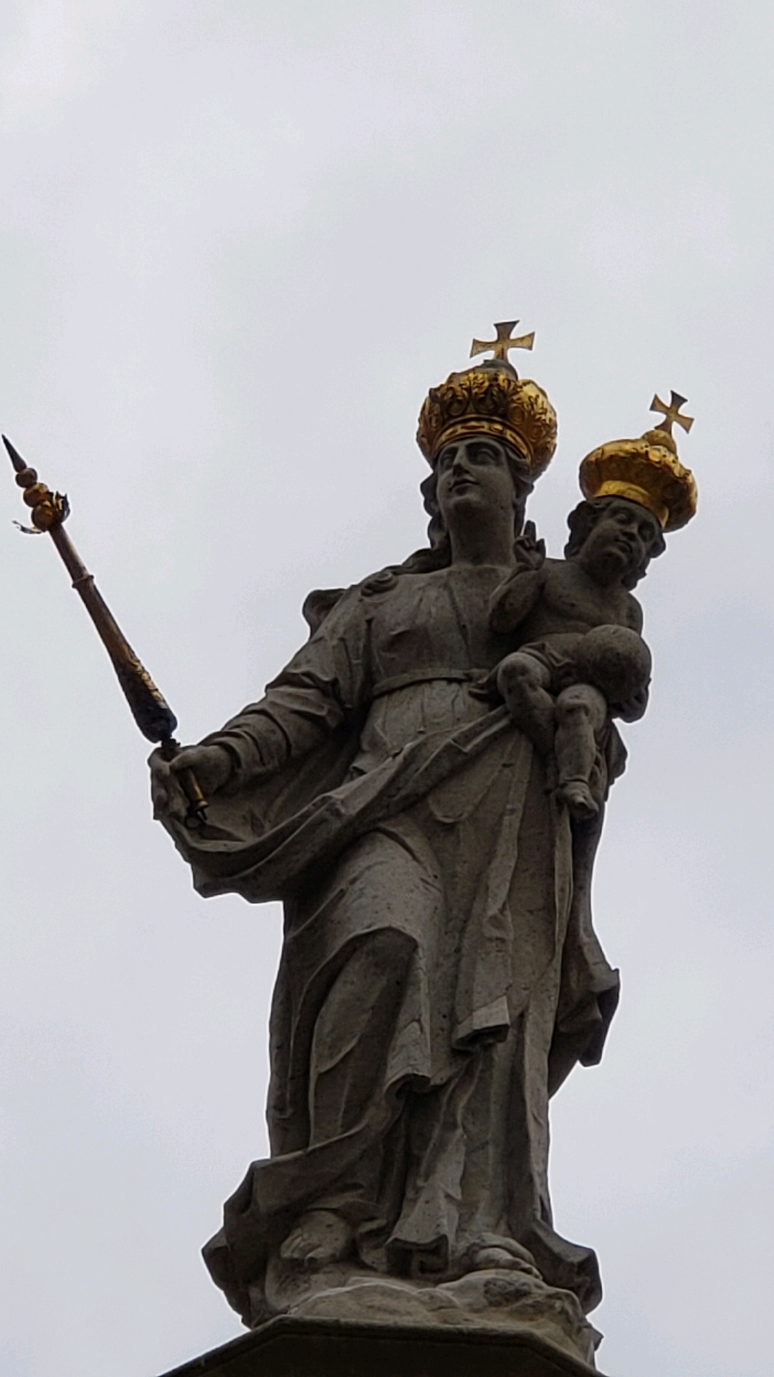 A stone statue of the Virgin Mary holding the infant Jesus is depicted from a low angle against a gray, overcast sky. Both figures wear ornate golden crowns topped with crosses. The Virgin Mary's crown is larger and more elaborate than Jesus's. She is dressed in flowing robes and holds a scepter in her left hand. The infant Jesus is cradled in her right arm, looking towards the viewer. The statue appears to be made of weathered stone, with some detailing visible in the drapery of Mary's dress and the faces of both figures. The statue is located in the city of Prešov, Slovakia.