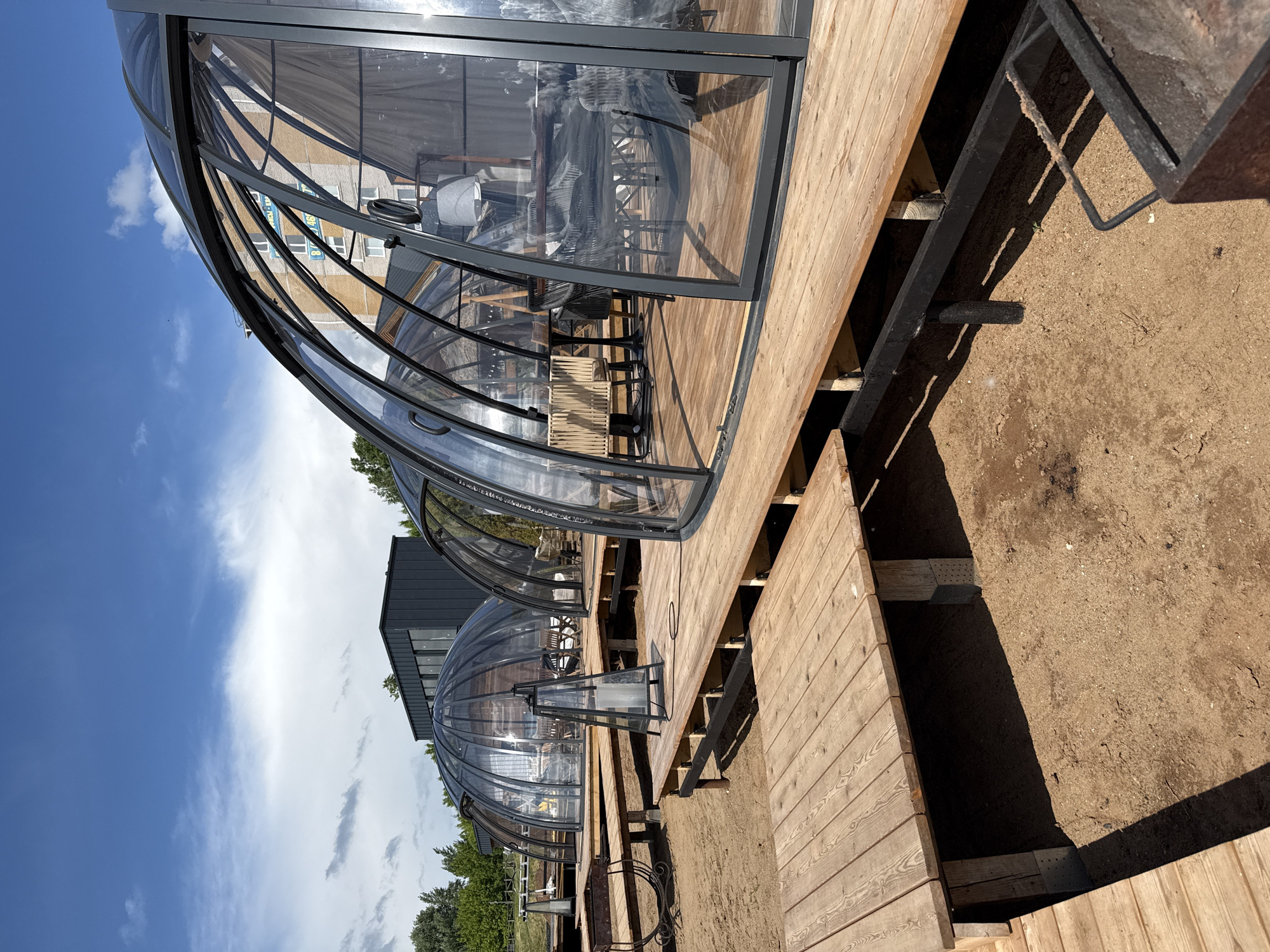 The image shows a row of clear, dome-shaped structures, possibly dining pods, located on a wooden deck area in Votkinsk, Russia. Each pod contains furniture, including tables and chairs, visible through the transparent walls. The deck is constructed of light-colored wood planks and appears to be elevated above a dirt or gravel ground surface. The sky is visible with a combination of blue sky and clouds. In the background, building is partially visible. The overall setting suggests an outdoor dining or recreational area.