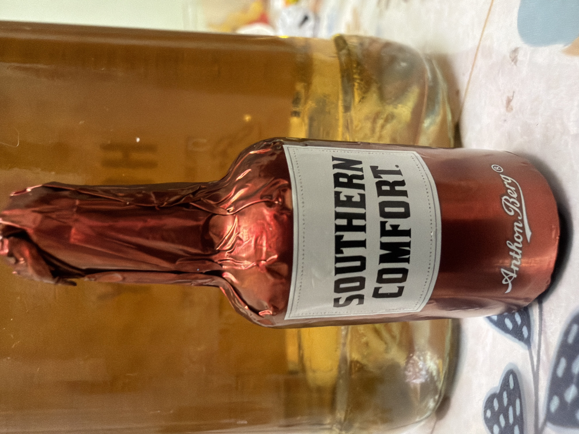 A close-up, eye-level shot shows a miniature bottle of Southern Comfort liquor, wrapped in shimmering red foil, standing in front of a larger glass bottle filled with amber-colored liquid. The miniature bottle has a white label with black lettering that reads "SOUTHERN COMFORT" in all caps. Below the label, also in white script, is the name "Anthon Berg". The background is a blurred amber liquid within a glass container, suggesting it might be another bottle or a large decanter. The scene appears to be indoors, with a patterned tablecloth beneath the bottles. The location is specified as Kyiv, Ukraine.