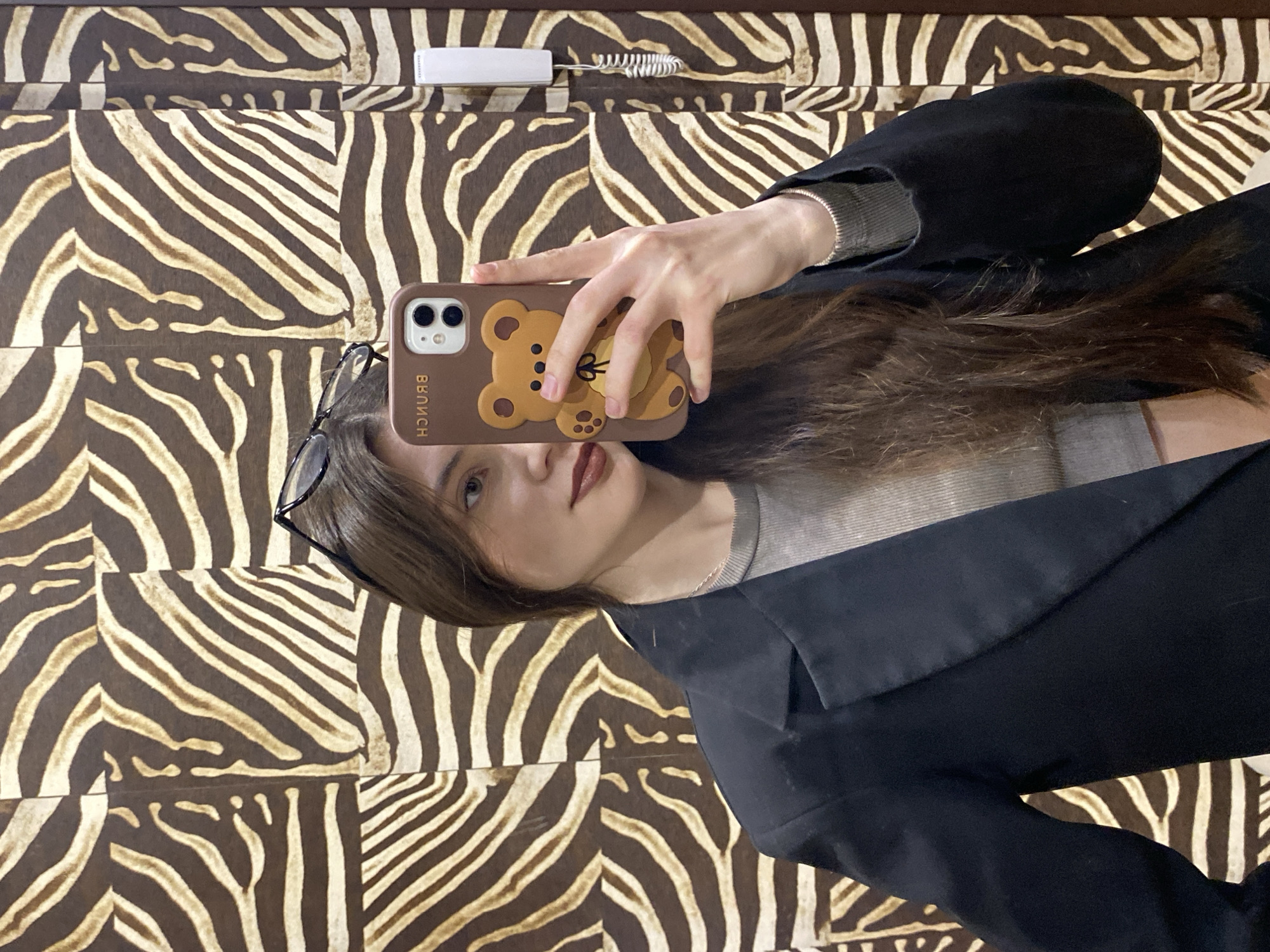 A selfie of a woman with brown hair and glasses is centered in the image. She holds up a phone with a brown case featuring a teddy bear design, partially obscuring her face. The woman wears a gray shirt under a black blazer.
The background consists of a zebra-striped pattern, likely wallpaper or fabric. At the top of the frame, a white telephone handset is visible.
The image does not contain sufficient information to determine its location in Votkinsk, Russia. The context provided is insufficient to give more information, such as subjects, objects, or actions.