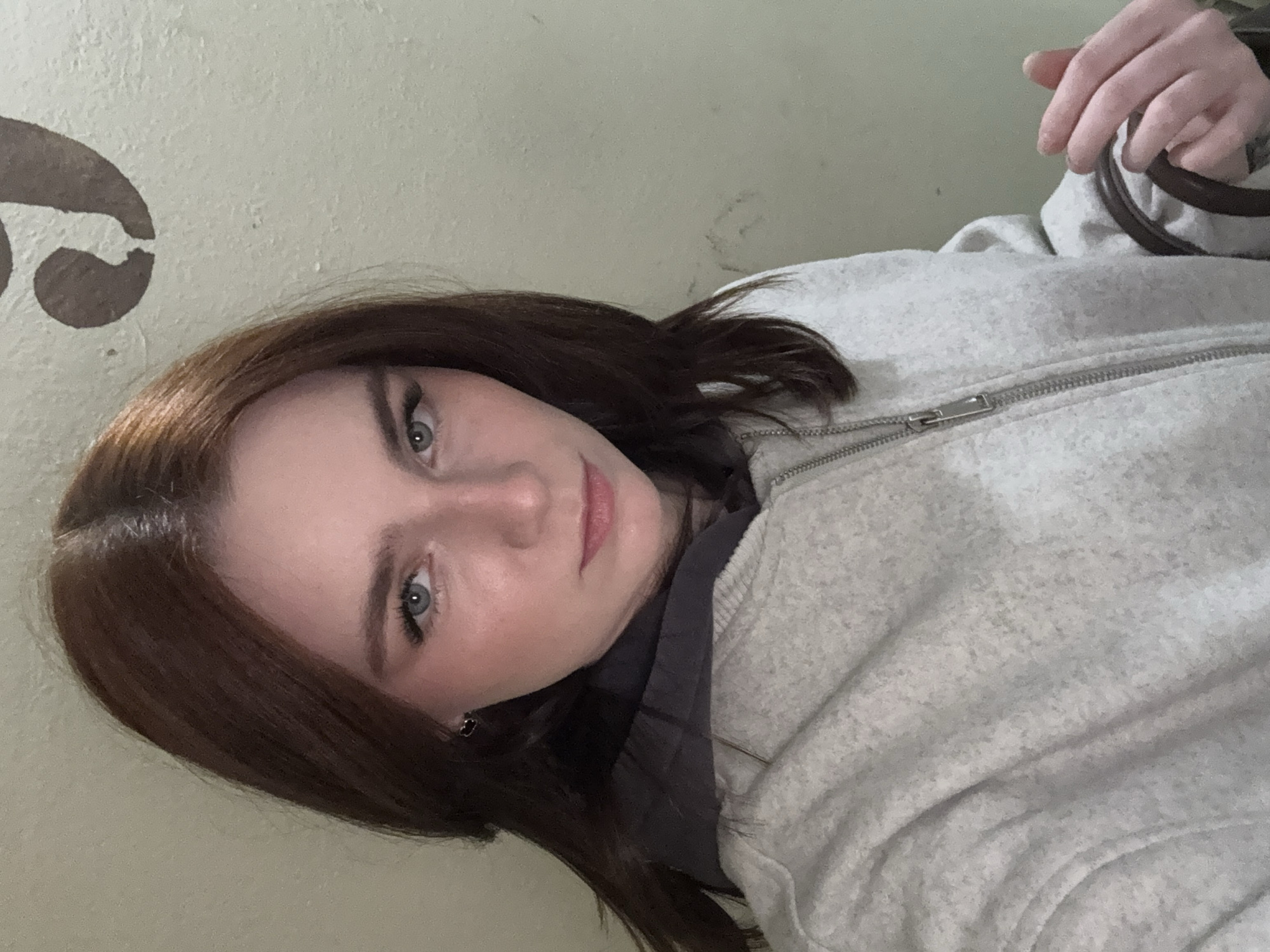 A fair-skinned individual with brown hair and blue eyes is seen in a medium close-up shot. The person is wearing a grey jacket with a zipper. A hand holding a dark, circular object is visible in the upper right corner. The background is a light, neutral color with an abstract brown design. The location is Unknown, Russia.